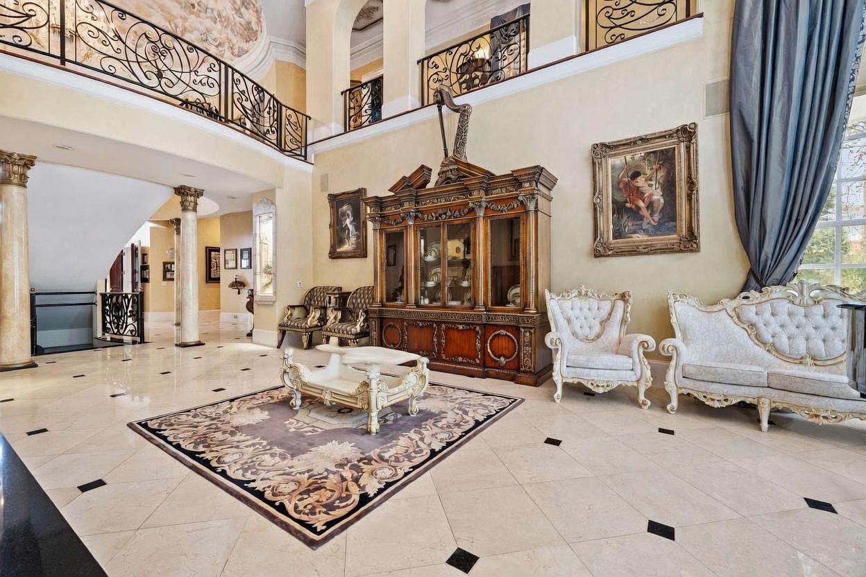 This grand, double-height living area features an opulent, traditional aesthetic with polished light-colored stone flooring accented by dark inlays. The space is anchored by a large, ornate wooden hutch topped with a harp, flanked by elegant white upholstered seating and a central coffee table on a patterned area rug. A sweeping wrought-iron balcony overlooks the room from the second level, enhancing the sense of scale and architectural luxury.