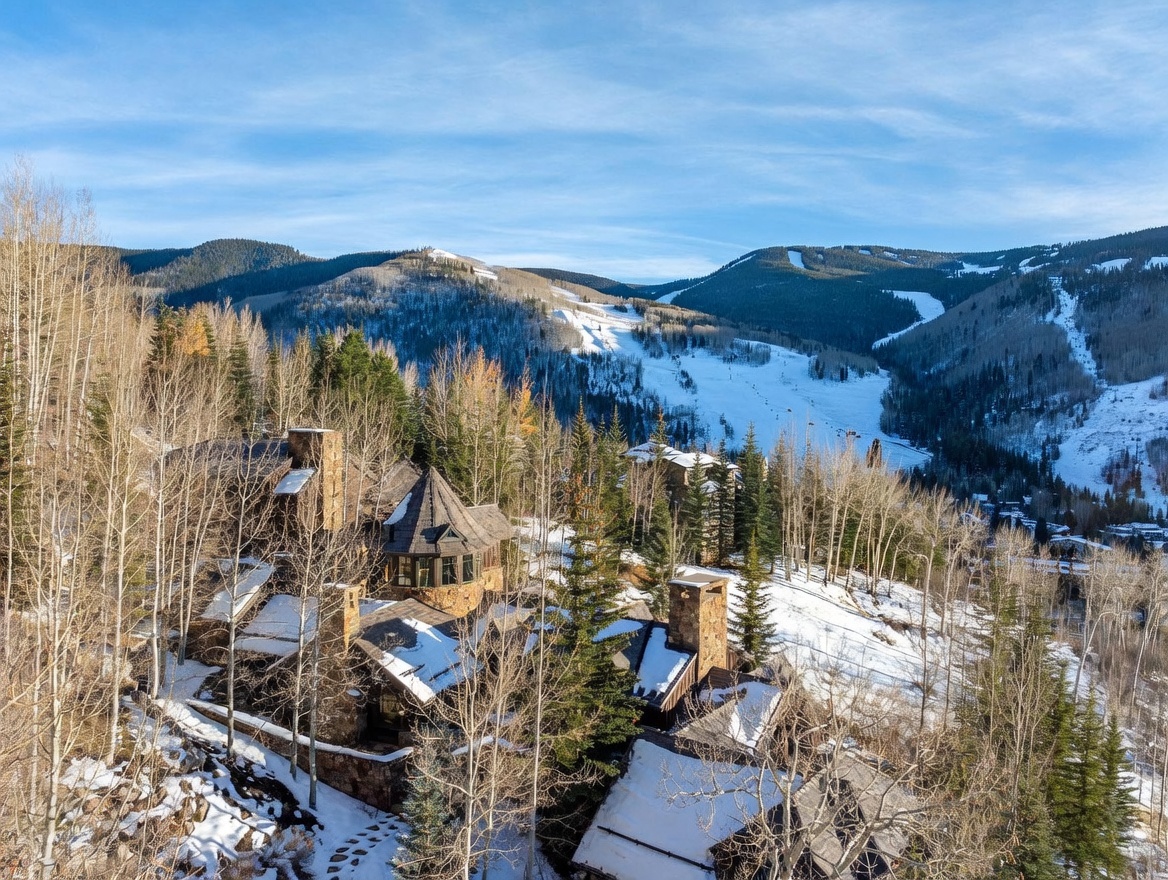 This elevated aerial perspective captures a sprawling mountain estate nestled within a dense forest of aspen and pine trees. The home features a complex, multi-gabled roofline with stone-clad chimney breasts and a distinctive turret, reflecting a rustic, alpine architectural style. In the background, a vast, snow-covered ski resort landscape stretches across the mountainside under a clear blue sky.