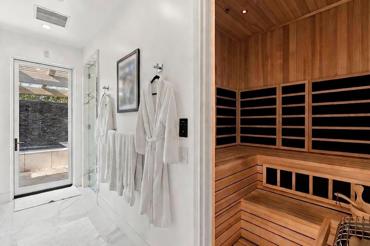 This is a luxurious primary bathroom featuring a sauna. The bathroom has white marble floors and walls, with a glass door leading to an outdoor area. The sauna is constructed of wood and has black panels. White robes hang on the wall, adding to the spa-like atmosphere.