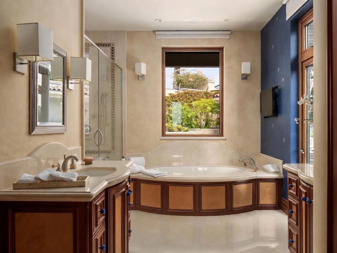 This luxurious primary bathroom features a central soaking tub set against a window with a garden view, flanked by dual vanities with stone countertops and dark wood cabinetry. The space is defined by a sophisticated color palette, combining warm beige walls with a striking deep blue accent wall and polished light-colored flooring. A glass-enclosed shower stall is positioned to the left, and the room is illuminated by wall-mounted sconces and recessed lighting, creating an elegant and serene atmosphere.