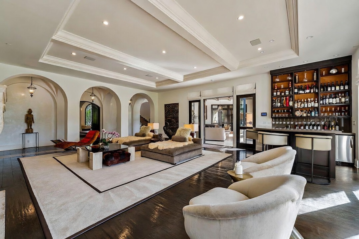 This expansive living room features dark hardwood floors, a neutral color palette, and a series of elegant arched doorways leading to adjacent spaces. The room is furnished with plush seating, including two curved armchairs in the foreground and a central lounge area with a low-profile coffee table. To the right, a built-in bar with extensive shelving and glass storage creates a sophisticated entertainment area, while recessed lighting and a coffered ceiling add architectural depth to the open-concept space.