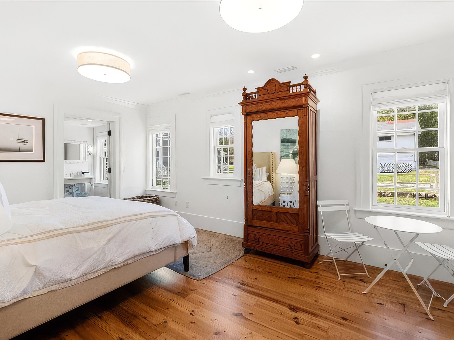This bright and airy bedroom features warm wood flooring and crisp white walls, creating a clean and inviting atmosphere. The room is furnished with a comfortable bed, a large antique-style wooden armoire with a mirrored door, and a small bistro-style table with two chairs near the window. Large windows allow for ample natural light, providing a pleasant view of the outdoor surroundings.