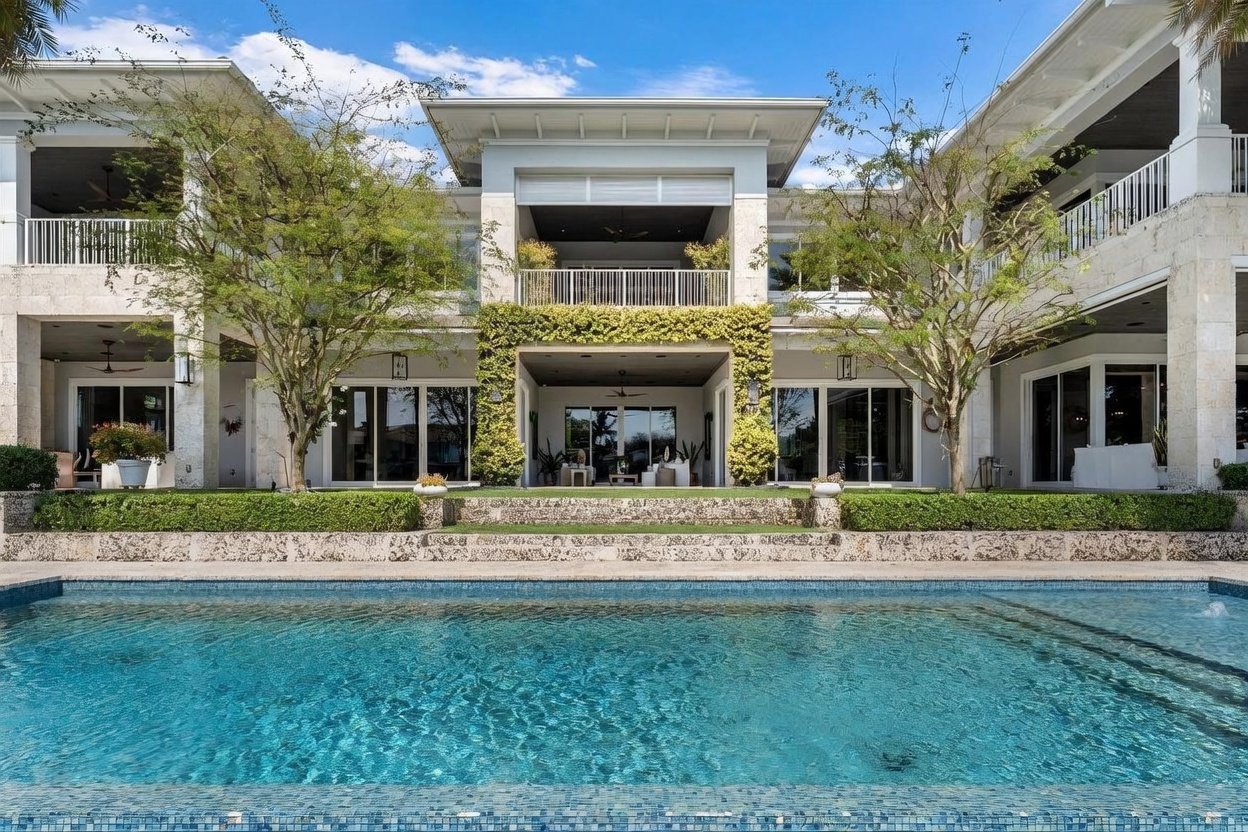 This expansive rear view of a luxury estate showcases a multi-level architectural design with a prominent swimming pool in the foreground. The home features stone-clad exterior walls, large glass sliding doors, and a covered balcony with a metal railing, all framed by lush green trees and manicured hedges. The scene is captured from a low, eye-level perspective across the pool, emphasizing the grand scale and resort-like atmosphere of the property.