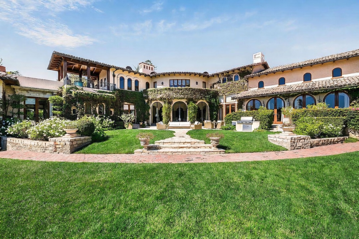 This expansive Mediterranean-style estate features a grand rear facade with arched doorways, climbing ivy, and a terracotta-tiled roof. A manicured lawn leads up to a stone-paved patio area, which is furnished with outdoor seating and a built-in grill station. The perspective is a wide, eye-level shot capturing the symmetry and architectural elegance of the home's rear elevation.