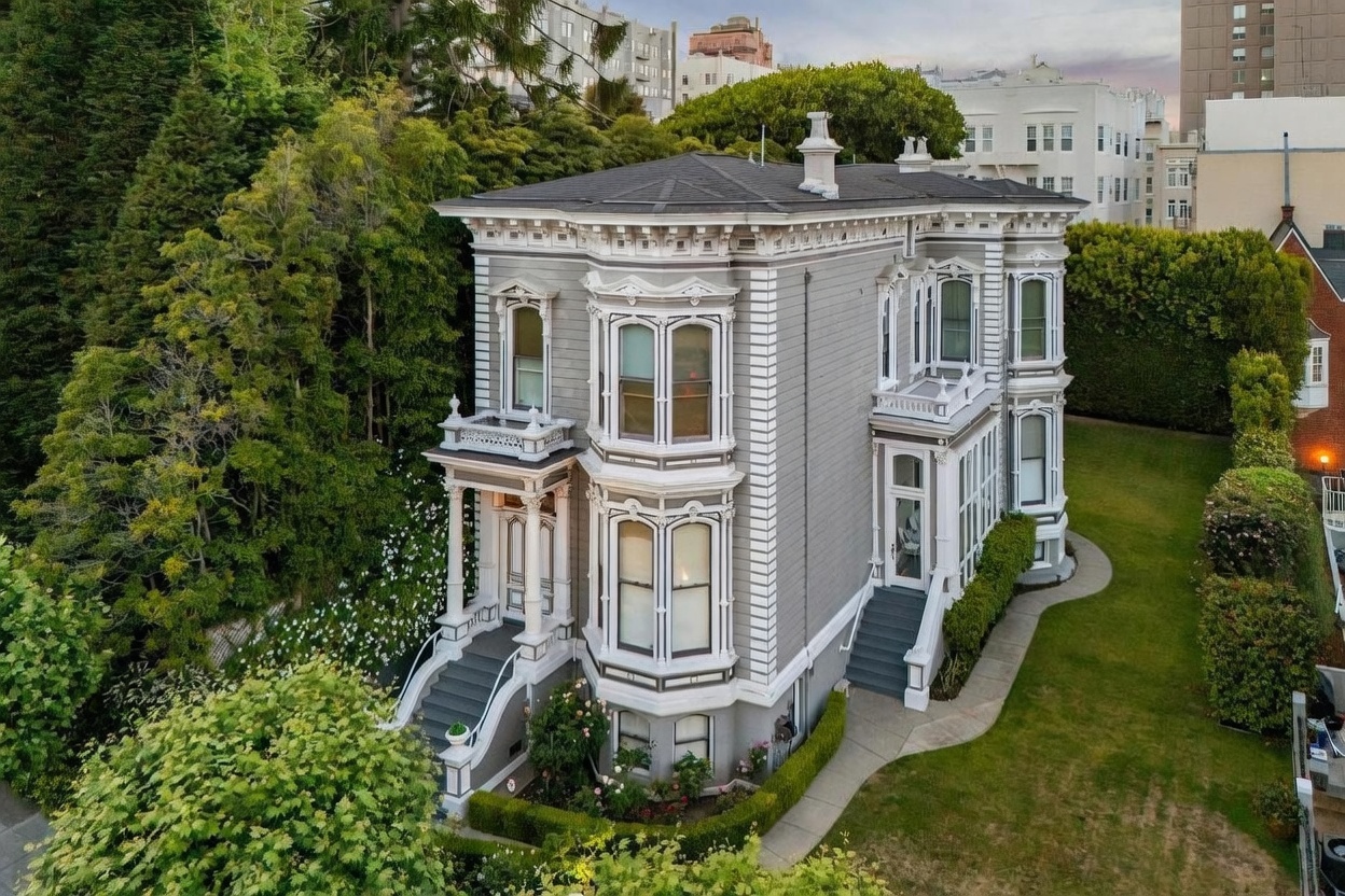 This elevated perspective captures a stately, multi-story Victorian-style home characterized by its intricate architectural detailing, grey wood siding, and white trim. The property features a prominent bay window structure, dual entry staircases, and a well-manicured lawn that wraps around the side of the building. Lush green trees frame the house, creating a sense of privacy and elegance in an urban setting.