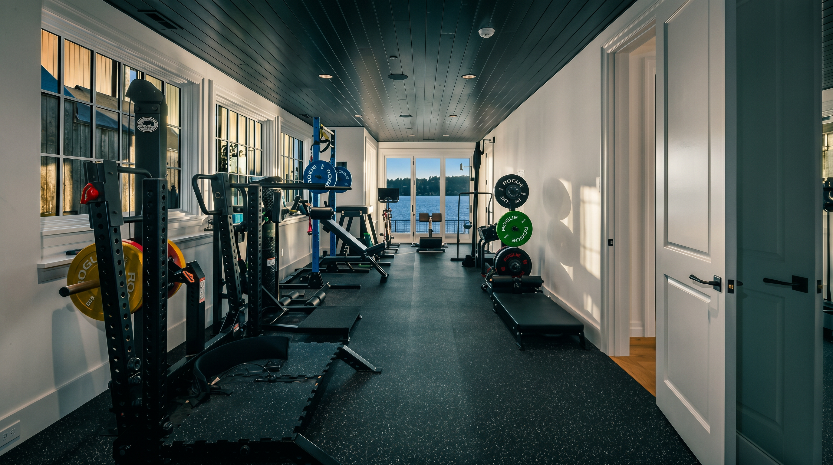 This professional-grade home gym features a long, narrow layout with dark, textured rubber flooring and a striking black-painted wood-plank ceiling. The space is equipped with a comprehensive Rogue fitness rack, weight plates, and various strength training equipment, all oriented toward a set of glass doors that provide a clear view of the water outside. The room is brightened by multiple windows along the side wall, creating an airy and motivating atmosphere for a workout.