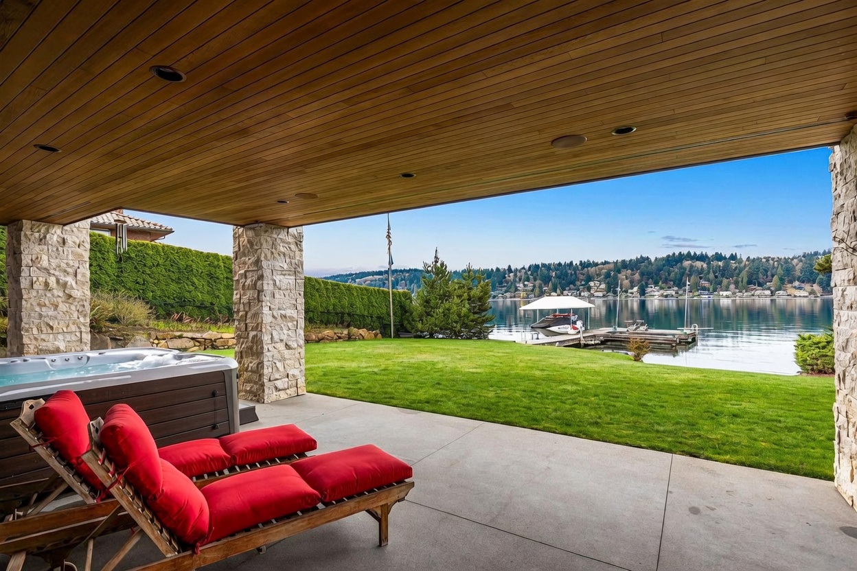 This covered patio offers a seamless transition to a lush green lawn and a serene waterfront view. The space features a built-in hot tub and two wooden lounge chairs with bright red cushions, all sheltered by a wood-paneled ceiling and supported by stone-clad pillars. The open-air design provides an expansive, unobstructed perspective of the lake, dock, and distant treeline.