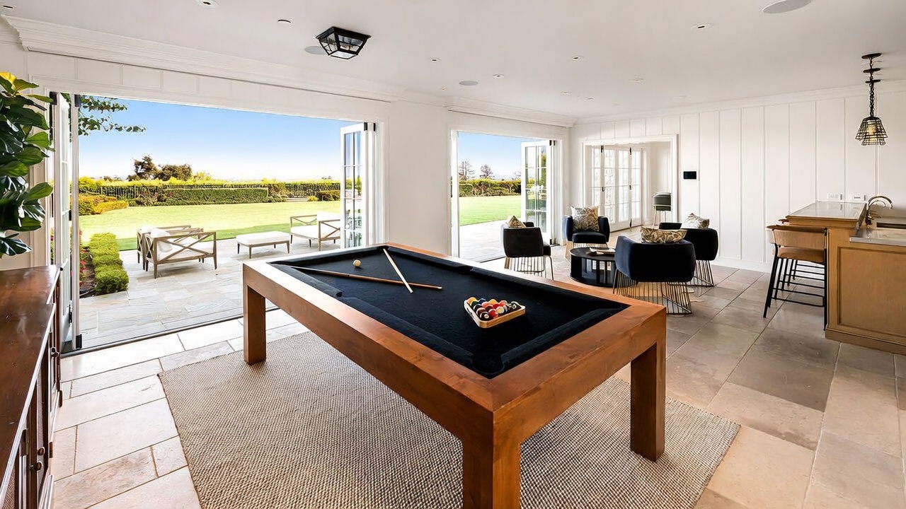 This spacious game room features a prominent wooden pool table as its centerpiece, set upon a textured area rug. The room seamlessly transitions to an outdoor patio through large, open glass doors, creating an airy and inviting atmosphere. To the right, a bar area with stools and a seating arrangement with dark armchairs provide additional space for relaxation and entertainment.