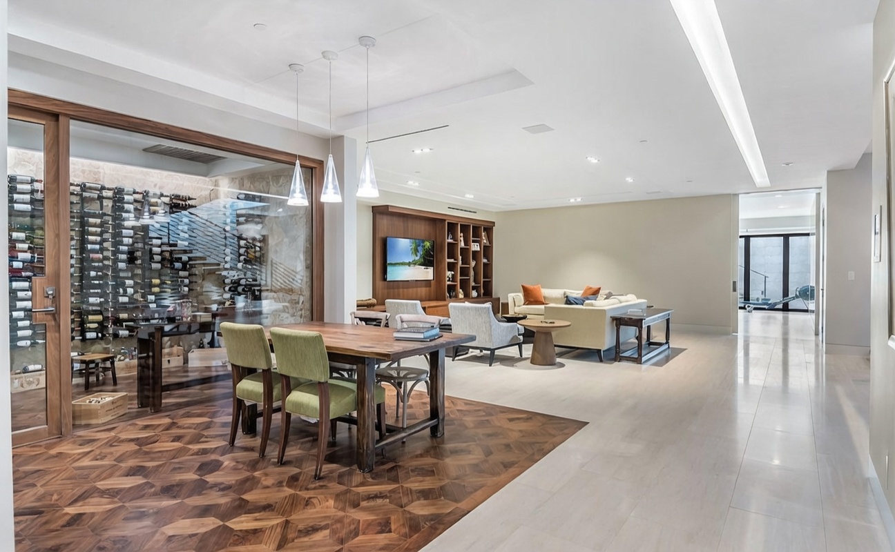 This open-concept living space features a dining area with a rustic wooden table and green upholstered chairs set upon a geometric patterned wood floor. Adjacent to the dining area is a glass-enclosed wine cellar, while the background reveals a spacious living room with a large sofa and a built-in media wall. The room is illuminated by modern pendant lights and recessed lighting, creating a bright and sophisticated atmosphere.