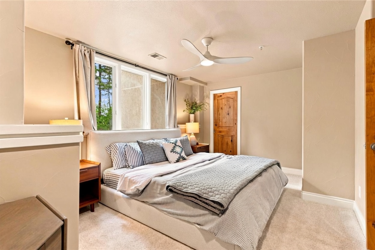 This cozy bedroom features a neutral color palette with a plush, light-colored upholstered bed frame as the central focus. The room is illuminated by a modern white ceiling fan and bedside lamps, while large windows with soft curtains allow natural light to brighten the space. A rustic wood door adds a touch of warmth and texture to the otherwise minimalist and serene design.