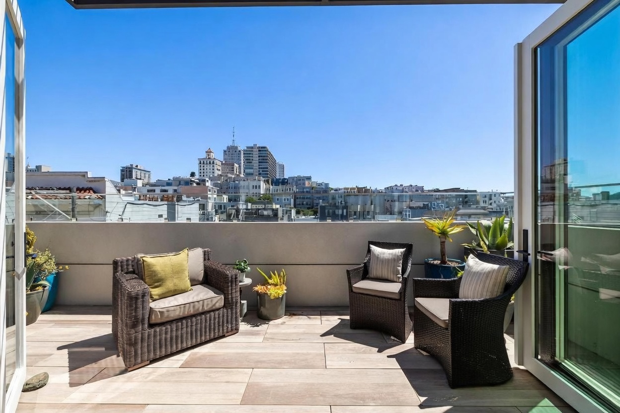 This modern balcony offers a panoramic view of the city skyline under a clear blue sky. The space is furnished with a wicker-style sofa and two matching armchairs, accented by potted succulents and greenery. Large glass doors open onto the tiled deck, creating a seamless transition between indoor and outdoor living areas.