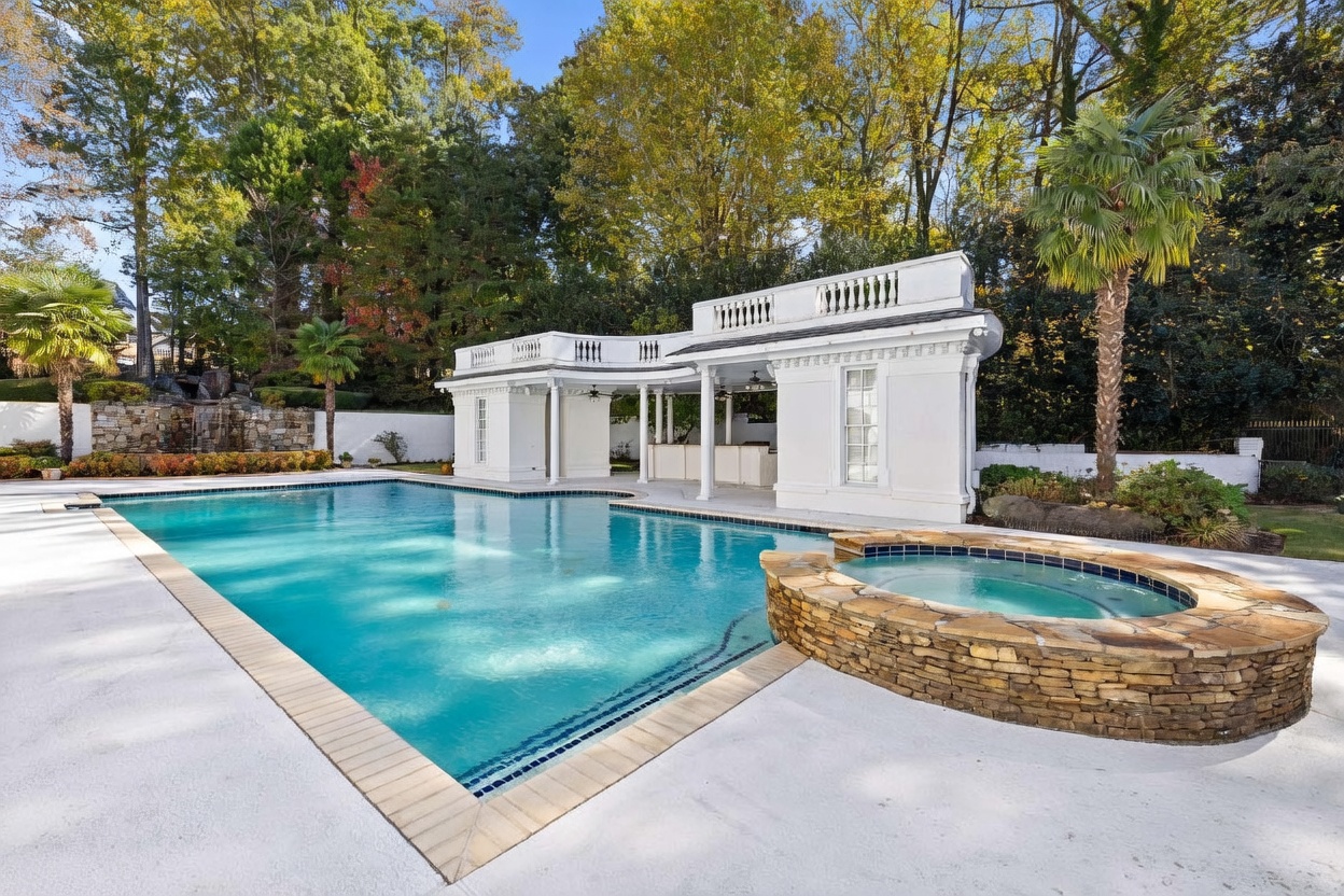 This luxurious backyard features a large, rectangular swimming pool paired with a circular, stone-clad spa. A white, classical-style pool house with columns and a balustrade roofline provides a shaded lounge area, while a stone waterfall feature adds a natural element to the background. The scene is framed by mature trees and palm trees, creating a private and resort-like atmosphere.