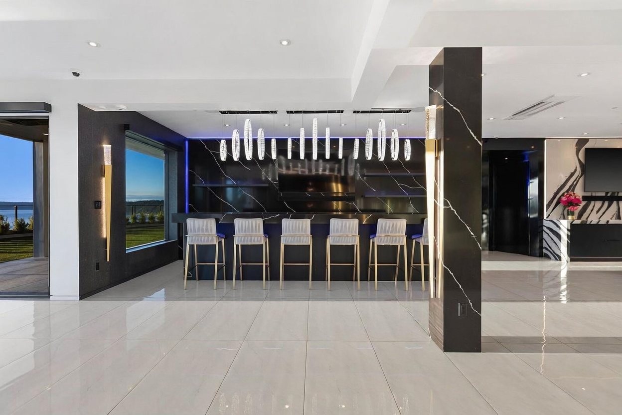 This modern, high-end bar area features a sleek black stone-clad wall with integrated lighting and a row of contemporary pendant lights hanging above a long counter. Six upholstered bar stools are arranged along the counter, which sits on a polished light-colored tile floor. To the left, large windows provide a view of the outdoors, while the overall aesthetic is sophisticated, dark, and luxurious.