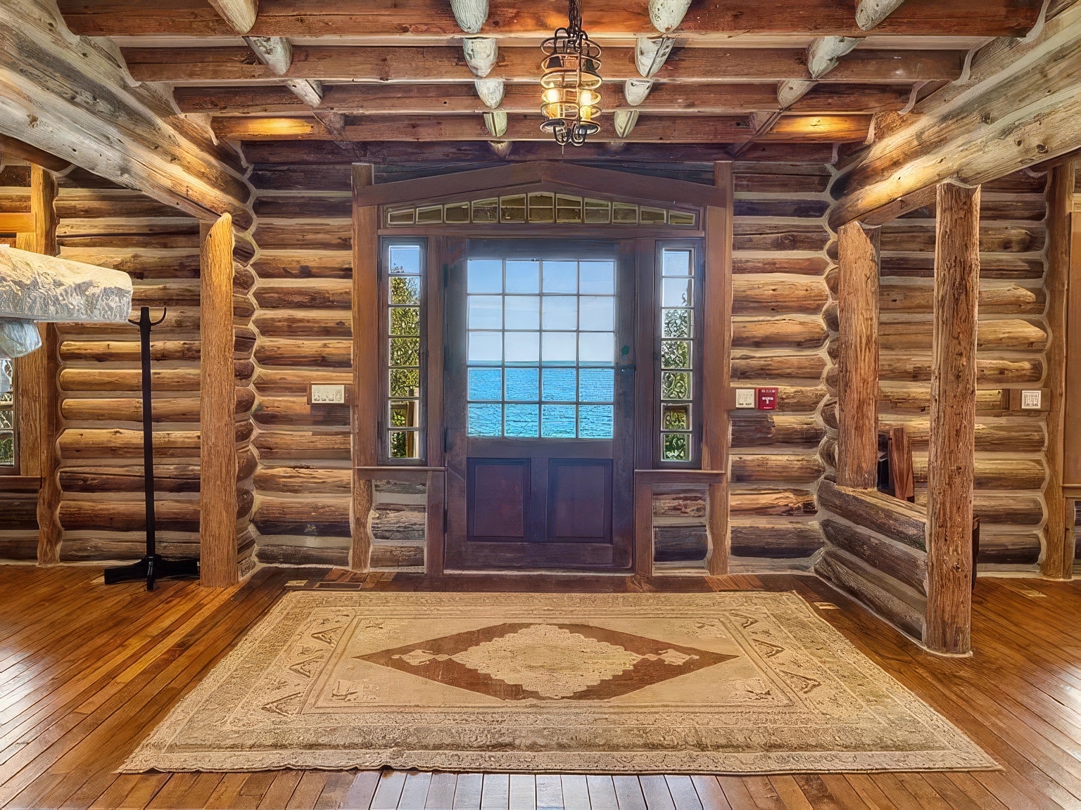 This rustic entryway features exposed log walls and a beamed ceiling, creating a warm and authentic cabin aesthetic. A central wooden door with glass panes provides a direct, scenic view of the ocean, while a large, patterned area rug anchors the polished wood floor. The space is illuminated by a decorative metal pendant light, emphasizing the natural textures and craftsmanship of the home.