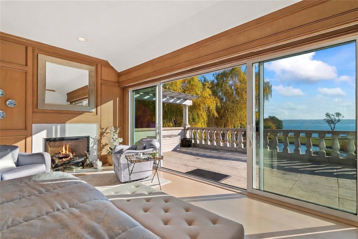 This luxurious primary bedroom features warm wood-paneled walls and a cozy fireplace with a stone surround. A large sliding glass door opens onto a spacious stone balcony, offering expansive views of the water and autumn trees. The room is furnished with a plush tufted bench in the foreground and comfortable armchairs, creating an elegant and serene atmosphere.