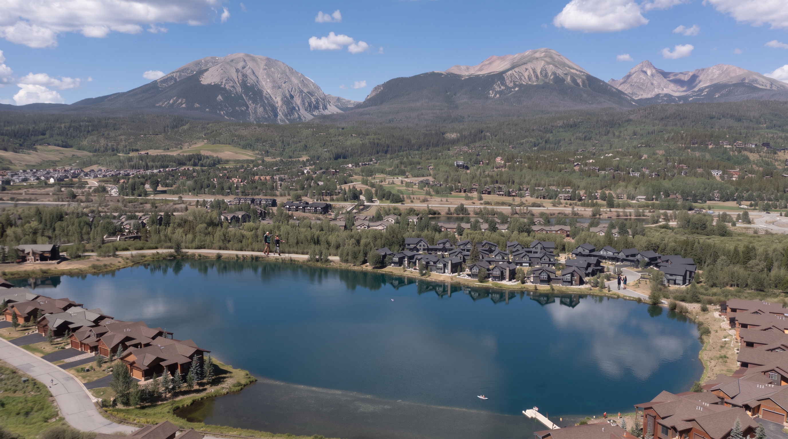 This stunning aerial view captures a luxury residential community nestled around a calm, reflective lake against a backdrop of majestic, snow-dusted mountains. The development features modern homes with dark, architectural finishes that blend seamlessly into the surrounding pine forests and natural landscape. The serene setting offers a perfect combination of outdoor living and sophisticated design, ideal for those seeking a tranquil mountain retreat.