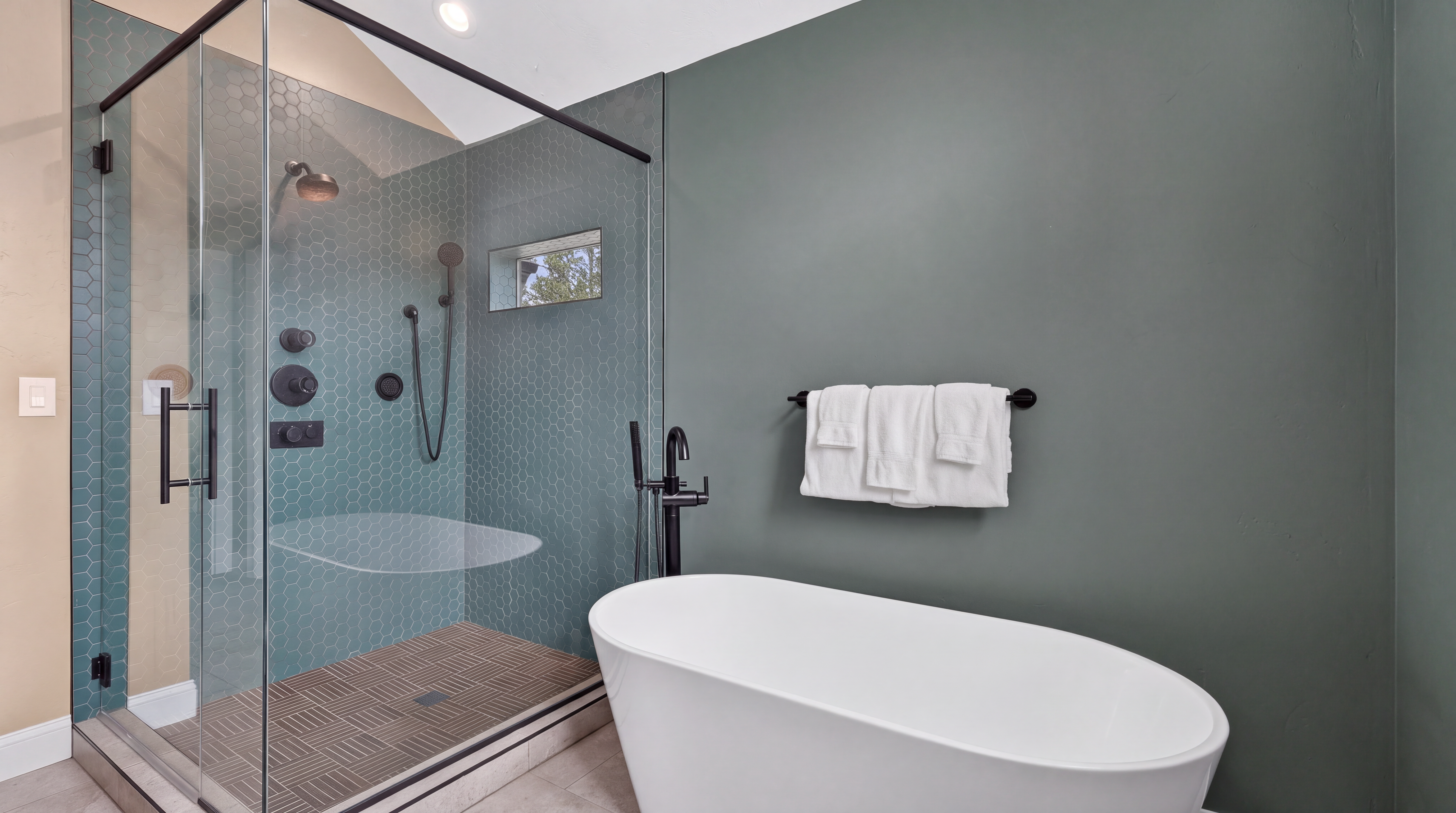 This modern bathroom features a sleek, freestanding white soaking tub positioned next to a walk-in shower enclosed by glass panels. The shower is accented with teal hexagonal wall tiles and a patterned tile floor, while the adjacent wall is painted in a deep, muted green. A black towel rack holds white towels, and black fixtures throughout provide a contemporary, high-contrast aesthetic.