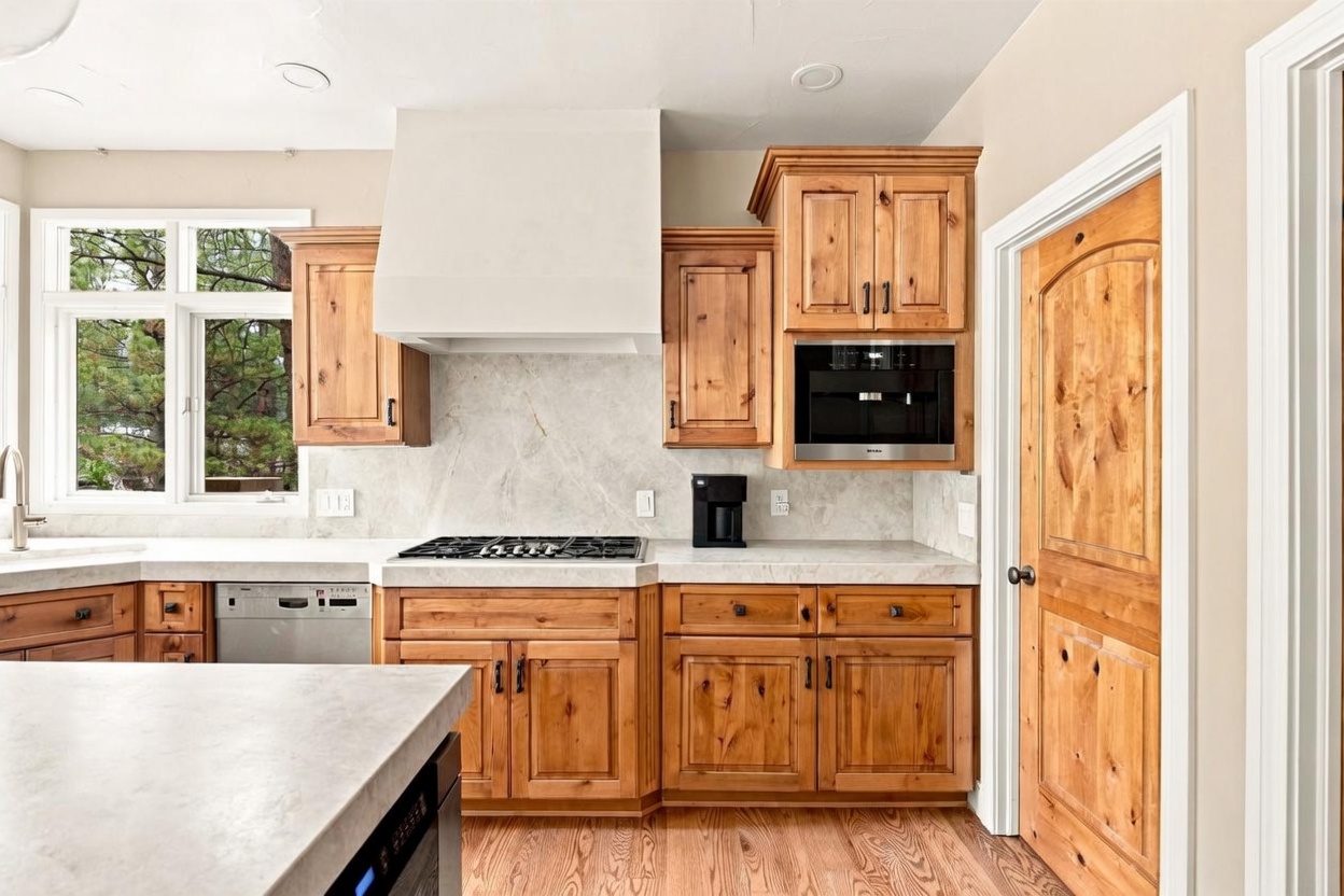 This kitchen features warm-toned wood cabinetry paired with light-colored stone countertops and a matching backsplash. The space is equipped with a gas cooktop, a built-in coffee machine, and a stainless steel dishwasher, all set against a neutral wall color. A large window above the sink provides natural light, while the overall design offers a rustic yet functional aesthetic.