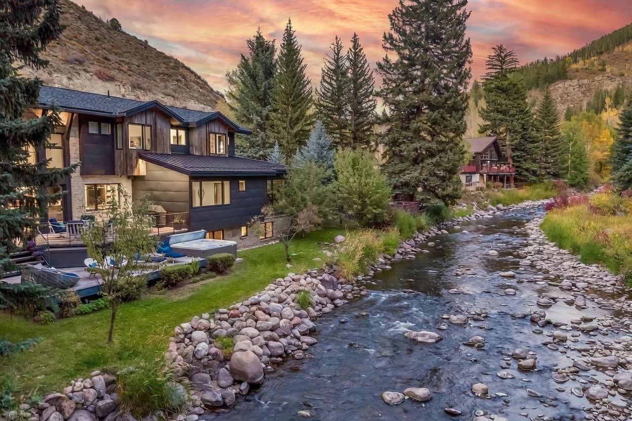 This elevated perspective captures the rear of a modern mountain home nestled along a rocky, flowing stream. The residence features a blend of dark wood siding and stone accents, with a multi-level deck that includes a private hot tub and outdoor seating areas. Surrounded by lush trees and a dramatic sunset sky, the property offers a serene and picturesque retreat in a natural landscape.