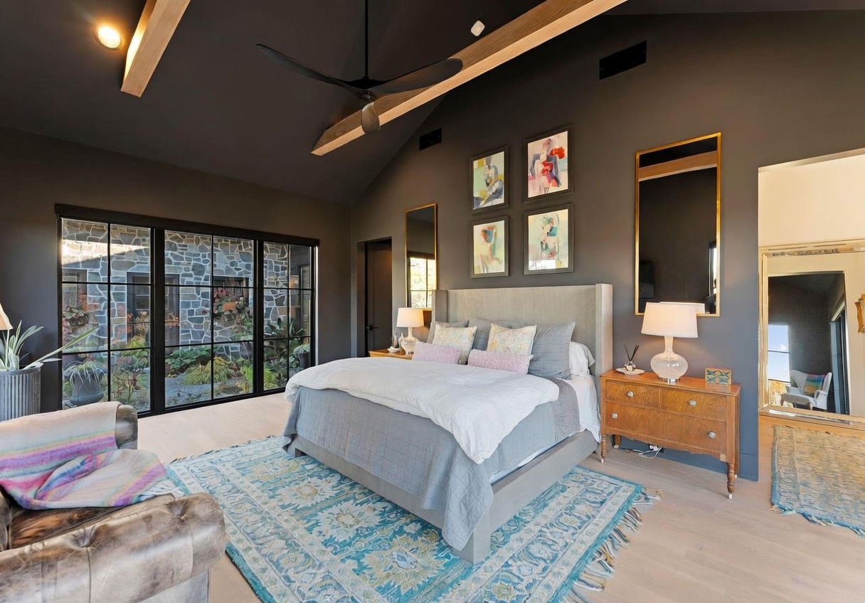 This spacious primary bedroom features a vaulted ceiling with exposed wooden beams and a dark, moody wall color that contrasts with the light-toned wood flooring. A large bed with a neutral upholstered headboard sits centered in the room, flanked by wooden nightstands and lamps, while a vibrant, patterned area rug adds a pop of color. Large, grid-paned windows provide a view of an exterior stone-walled courtyard, and a tall mirror leans against the wall, enhancing the room's airy and sophisticated atmosphere.