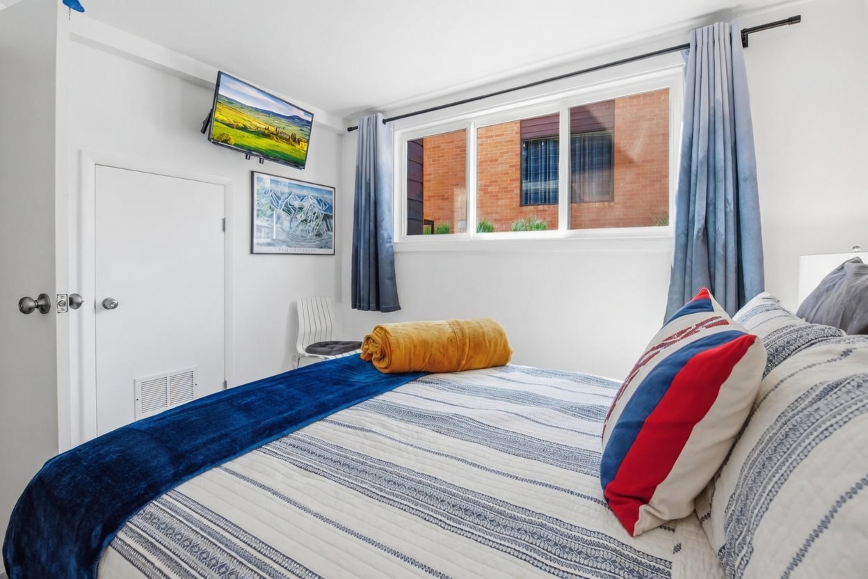 This cozy bedroom features a bed dressed in a blue and white striped quilt, accented by a vibrant red, white, and blue decorative pillow and a folded mustard-yellow throw blanket. A wall-mounted television is positioned above a framed print of a ski map, while a simple white chair sits in the corner near a window with blue ombre curtains. The room is bright and minimalist, offering a comfortable and functional space with a view of a brick building exterior.