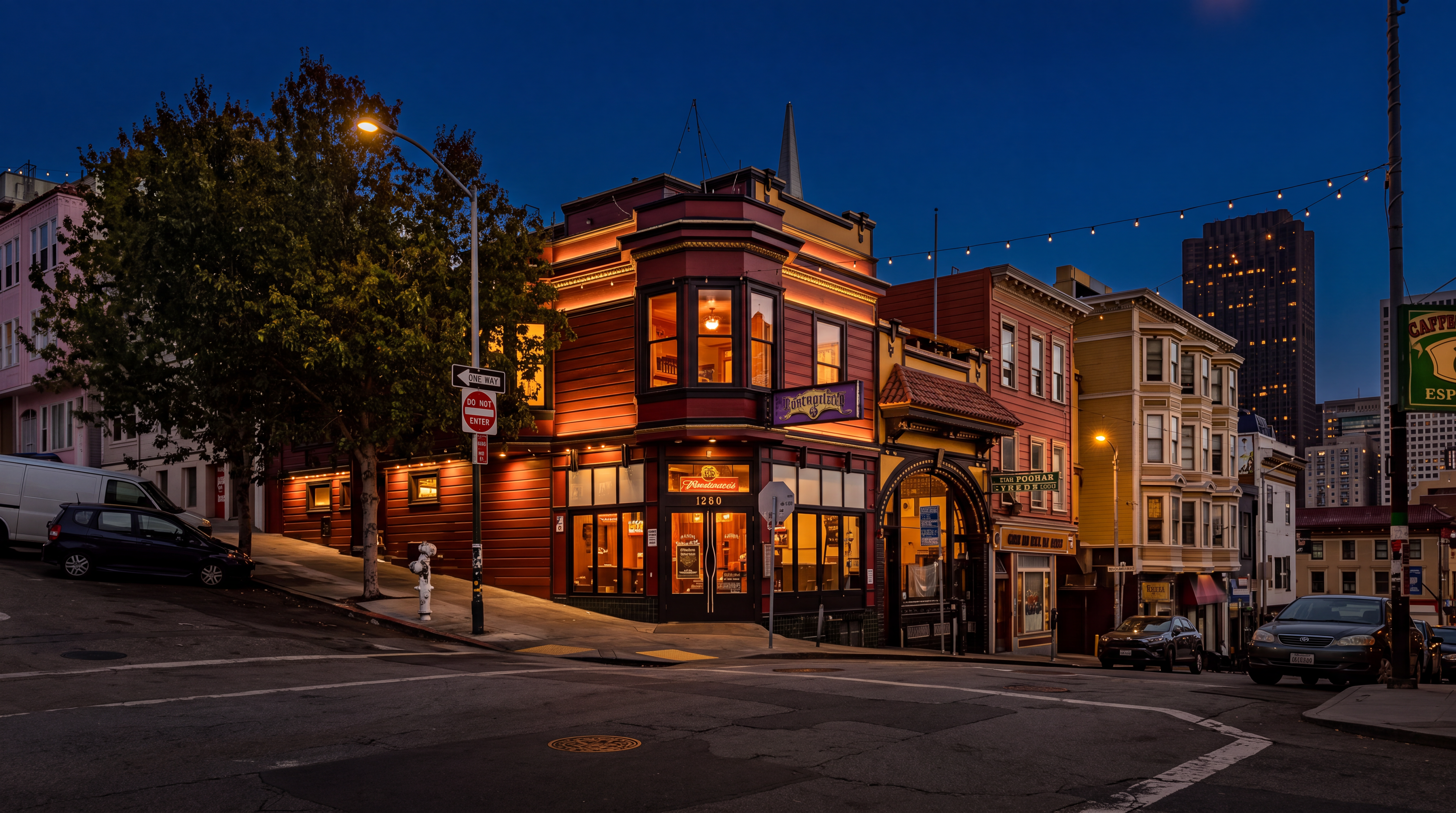This stunning night-time shot showcases a historic, corner-lot building in a vibrant San Francisco neighborhood. The structure features rich, red-toned wood siding, signature bay windows, and a classic architectural arch, all beautifully illuminated against a twilight sky. The setting includes a busy street intersection, neighboring multi-story city buildings, and overhead string lights, creating an inviting and quintessential urban atmosphere.