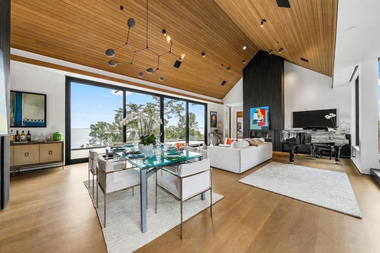 This expansive open-concept living and dining area features a dramatic vaulted wood-paneled ceiling and large glass sliding doors that frame a scenic water view. The space is anchored by a tall, dark-paneled fireplace surround and includes a grand piano, a contemporary dining set, and a plush white sofa. The light wood flooring and neutral color palette create a sophisticated, airy atmosphere perfect for entertaining.