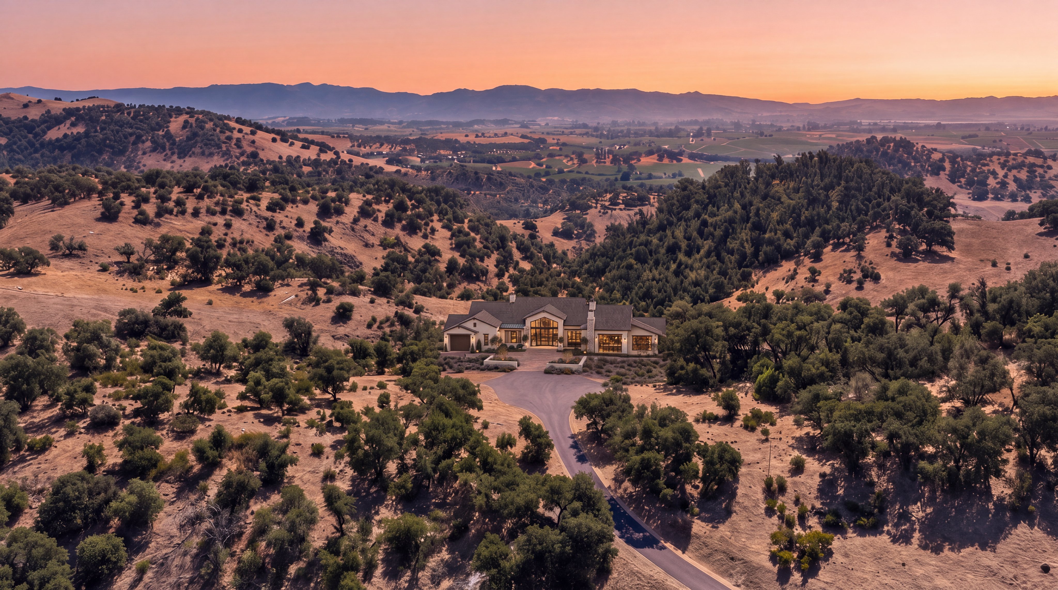 This stunning aerial shot captures a large, luxurious estate nestled within a vast, rolling landscape of golden hills and mature oak trees during a picturesque sunset. The home features a grand architectural design with warm interior lighting glowing through expansive windows, and a paved driveway leading up to its welcoming entrance. The scene provides an impressive sense of scale, privacy, and serene natural beauty, showcasing the property's secluded and elevated positioning.