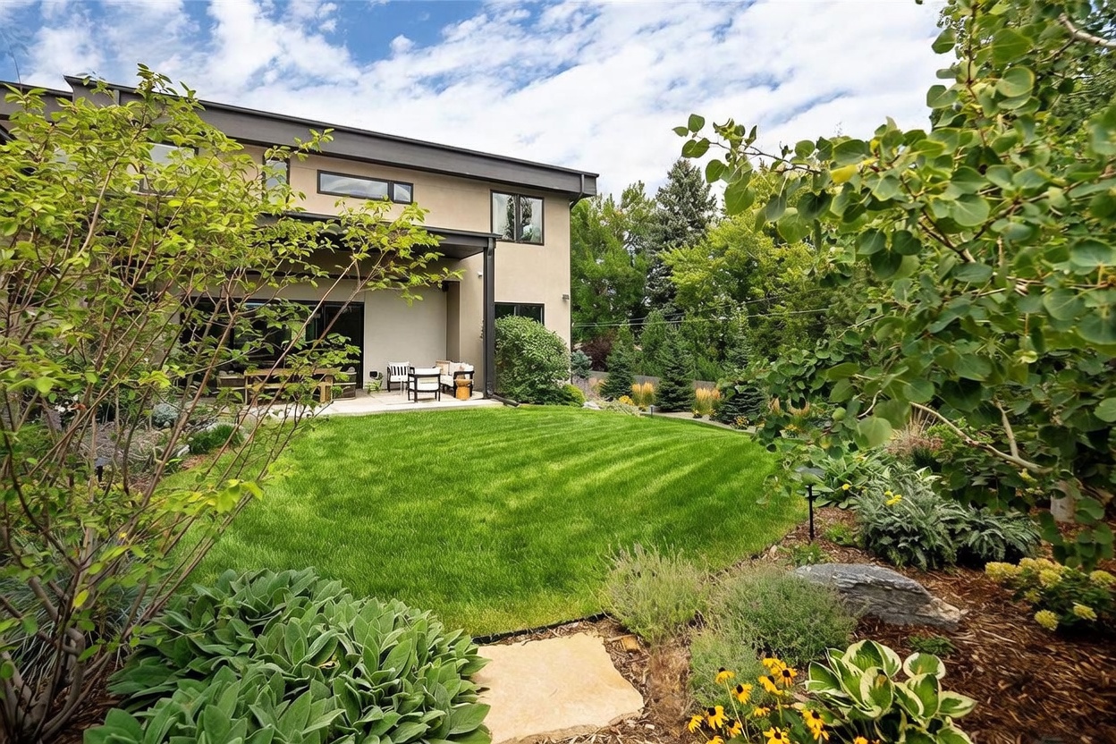 This rear view of a modern home showcases a well-manicured lawn and a covered patio area furnished with outdoor seating. The exterior features a neutral-toned facade with clean lines and large windows, surrounded by lush landscaping including mature trees and flowering plants. The perspective is from a garden path, looking toward the house, creating a serene and inviting backyard atmosphere.