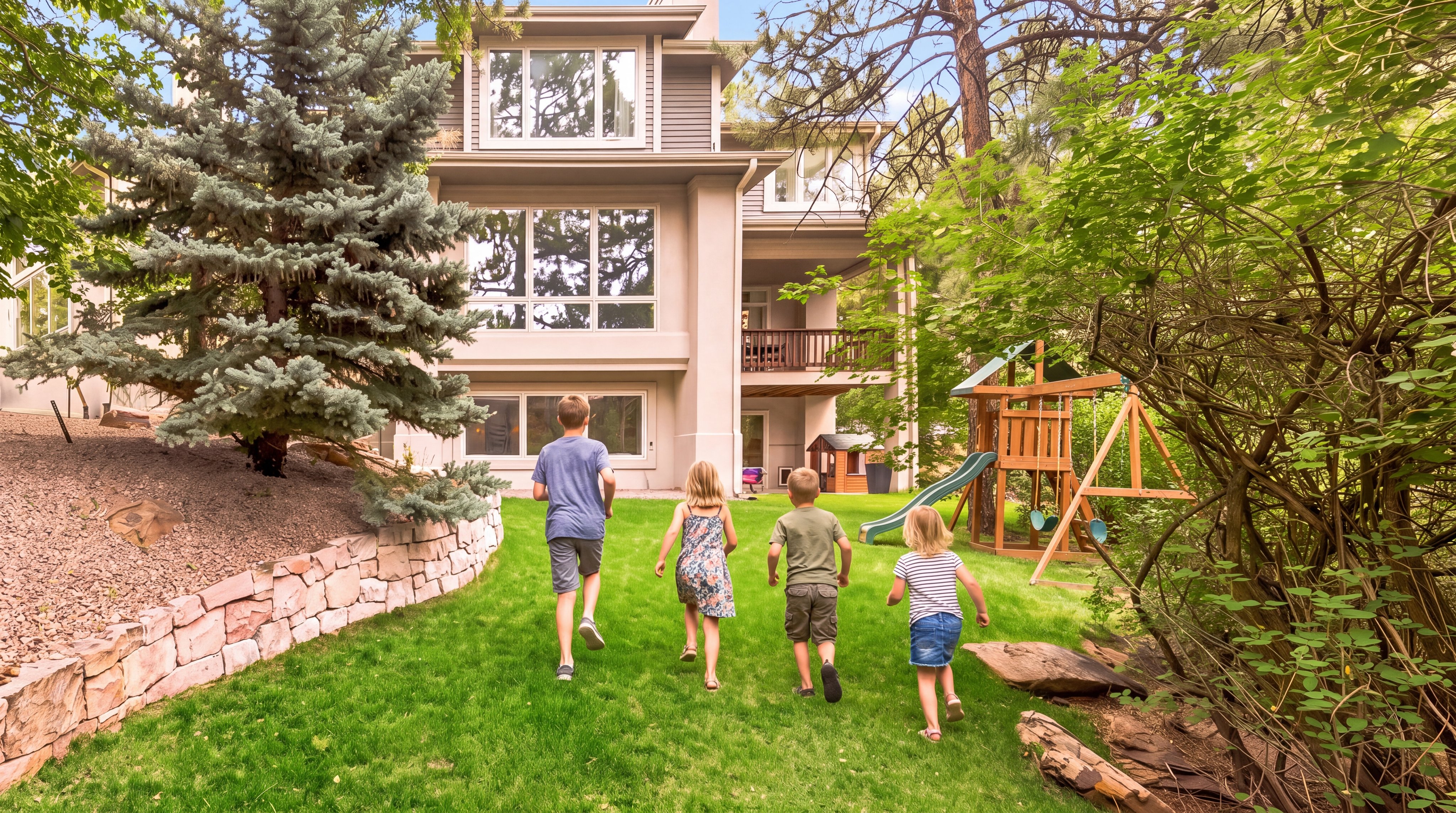 This spacious, lush green backyard offers a family-friendly setting, featuring a well-manicured lawn and a wooden swing set structure. The rear view of the multistory home showcases large windows, a raised wooden deck, and a natural stone retaining wall, blending comfortable residential living with a peaceful, wooded environment.
