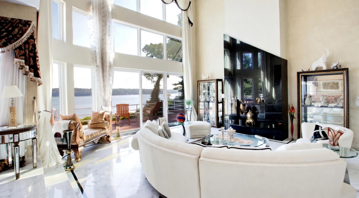 This grand, double-height living room showcases opulent, traditional decor with massive floor-to-ceiling windows offering expansive waterfront views. The space features elegant white marble flooring, a striking black marble-faced fireplace, and luxurious curved cream-colored seating, creating a sophisticated and airy atmosphere. The room is further accented by ornate valances, a crystal chandelier, and formal display cabinets, reflecting a high-end, luxury aesthetic.