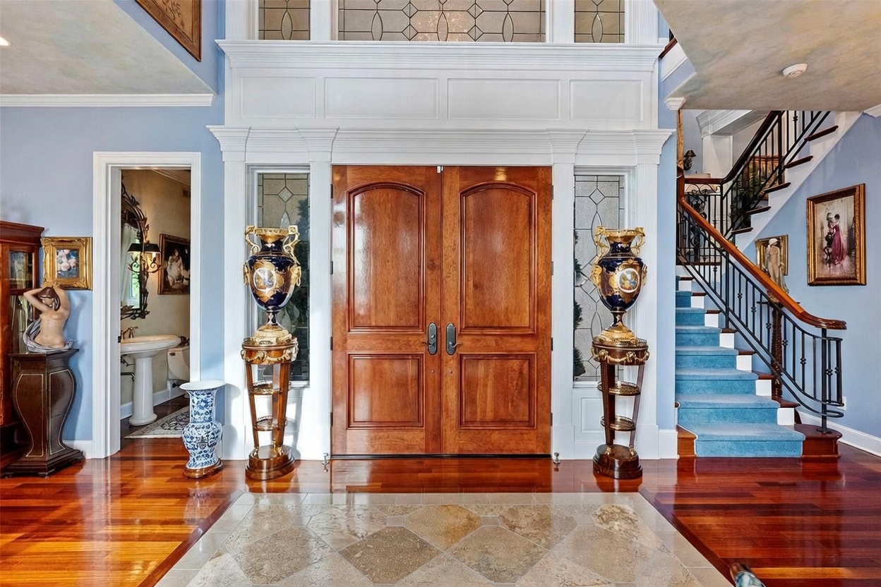 This grand foyer features a double-door entrance flanked by decorative urns on pedestals and sidelight windows. A staircase with a dark metal railing and blue carpeting ascends to the upper level, while a stone-tiled inlay transitions into polished wood flooring. The space is accented by ornate artwork and a glimpse into an adjacent powder room, creating a formal and elegant first impression.