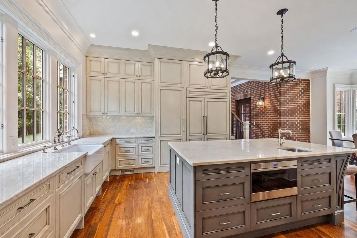 This elegant kitchen features a large central island with a dark base and a light stone countertop, complemented by light-toned perimeter cabinetry and a farmhouse-style sink. The space is illuminated by two decorative pendant lights hanging above the island, while a brick wall adds a rustic texture to the background. The warm wood flooring and large windows create an inviting and bright atmosphere, perfect for both cooking and entertaining.