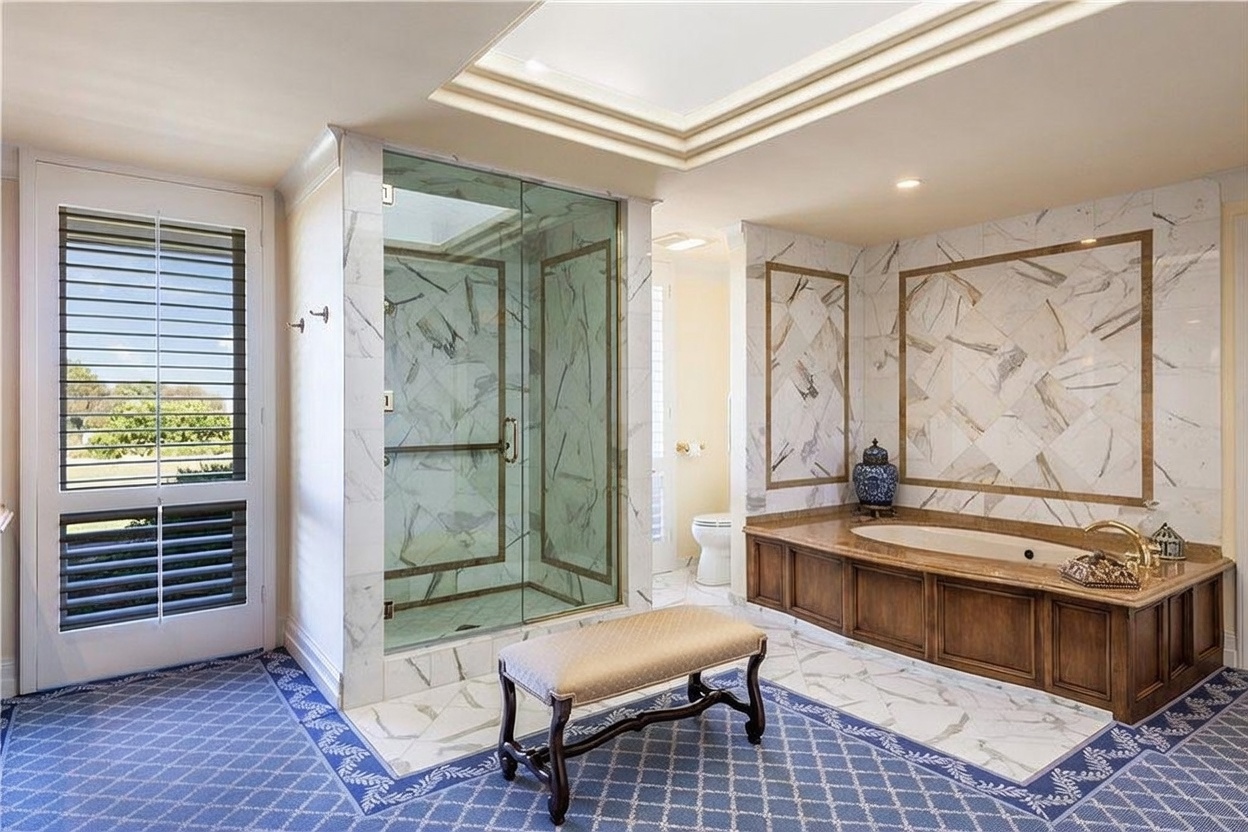 This spacious primary bathroom features a large soaking tub set within a wood-paneled surround, accented by decorative stone wall tiles. A glass-enclosed shower with stone tiling stands centrally, while a cushioned bench sits on the patterned blue carpet in the foreground. The room is brightened by a skylight and a door with glass panes that leads to an outdoor area.