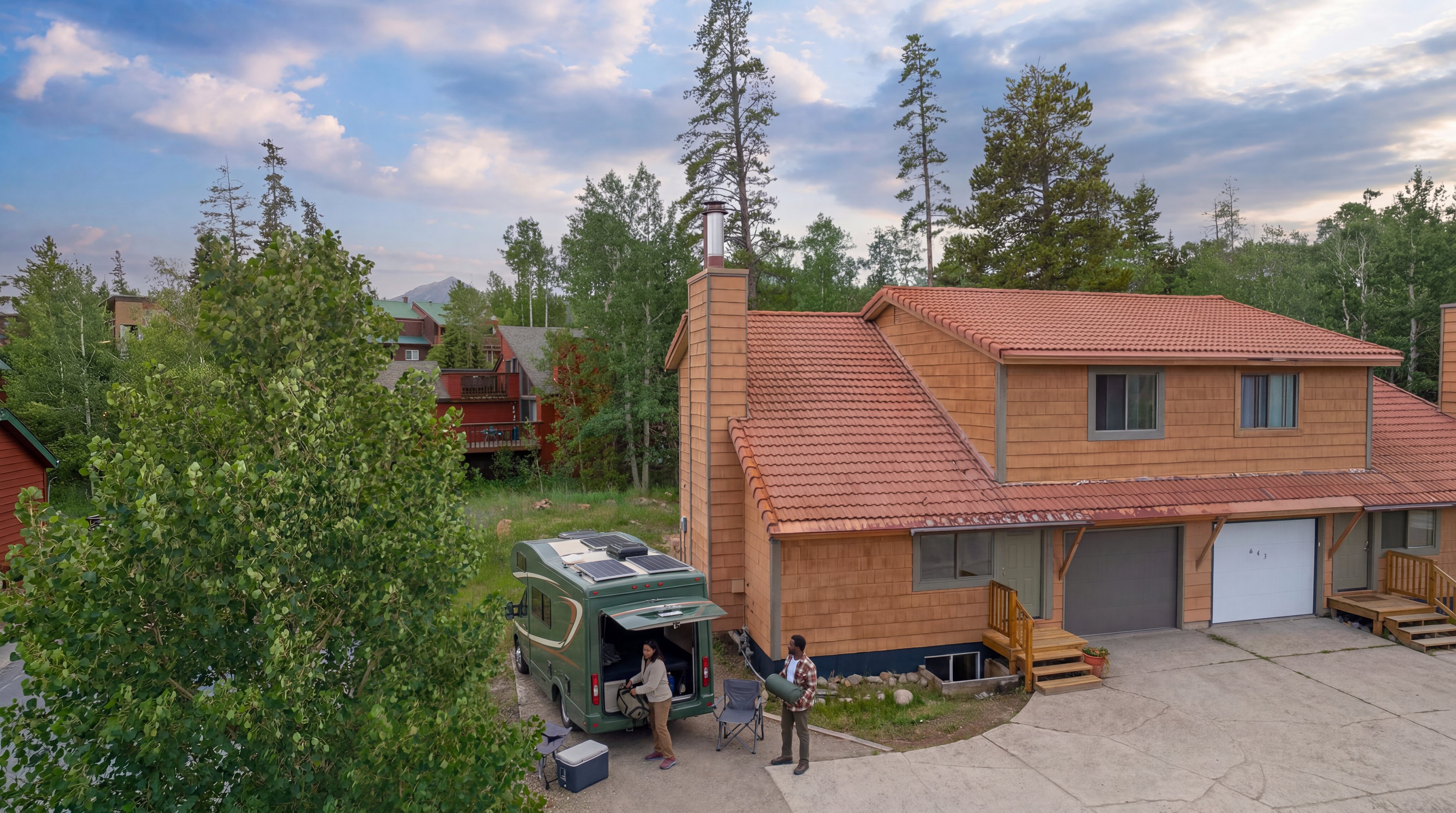 This elevated view captures a two-story residential home with textured wood-shingle siding and a prominent tile roof, nestled in a scenic, wooded environment. The exterior includes an attached garage and a multi-level deck, while a van is parked in the driveway with people nearby, suggesting a relaxed, outdoor lifestyle. The property's integration with the natural landscape and mountainous backdrop adds significant aesthetic appeal.