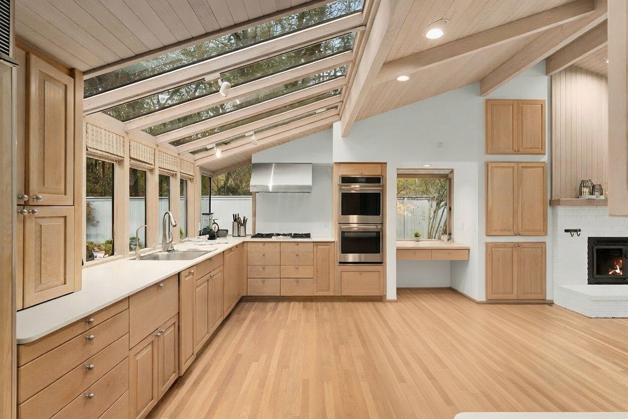 This bright, modern kitchen features light wood cabinetry and a large central island with a white countertop and integrated sink. A striking sloped glass ceiling allows abundant natural light to flood the space, highlighting the clean lines and open layout. The kitchen is equipped with stainless steel appliances, including a double wall oven and a gas cooktop, while a fireplace with a white brick surround adds a cozy touch to the adjacent living area.