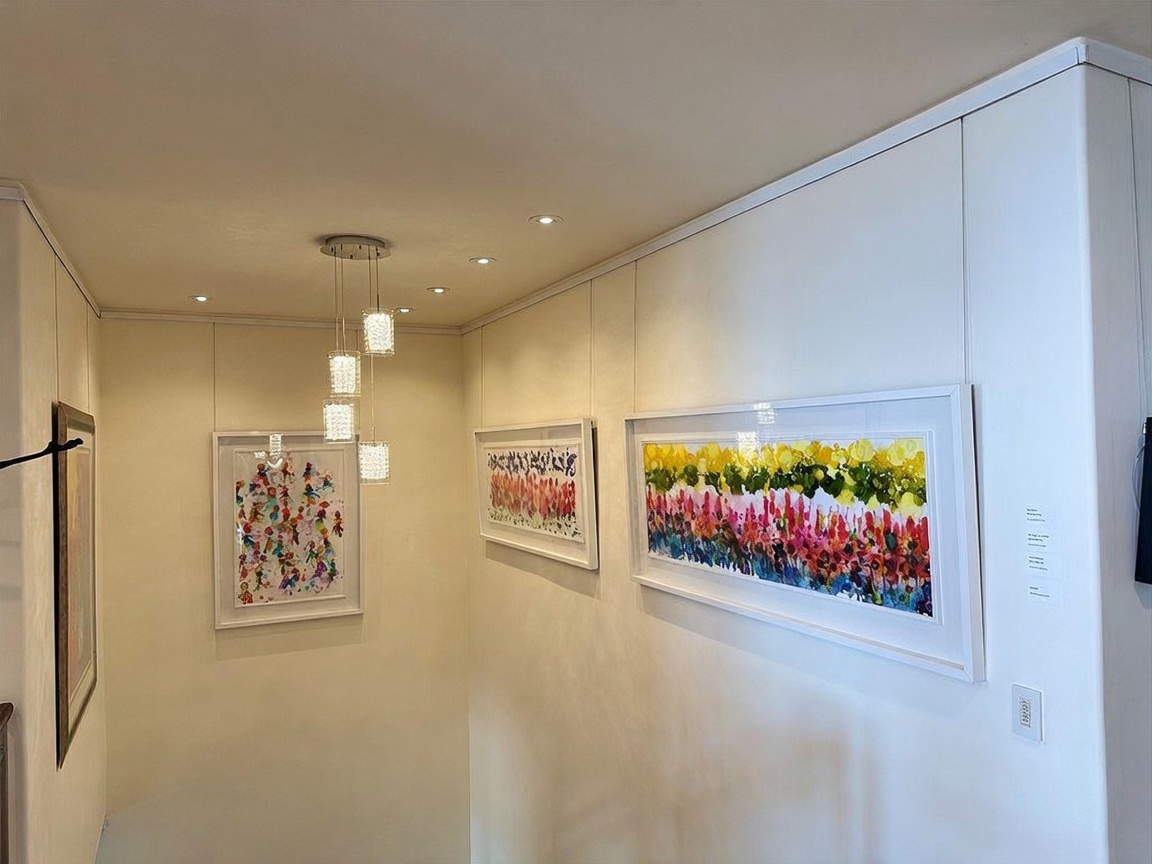 This bright hallway features a series of vibrant, abstract art pieces displayed on clean, neutral walls. A modern, multi-pendant light fixture hangs from the ceiling, casting a warm glow over the space, while recessed lighting provides additional illumination. The perspective looks down a staircase, highlighting the gallery-like atmosphere of the home's transition area.