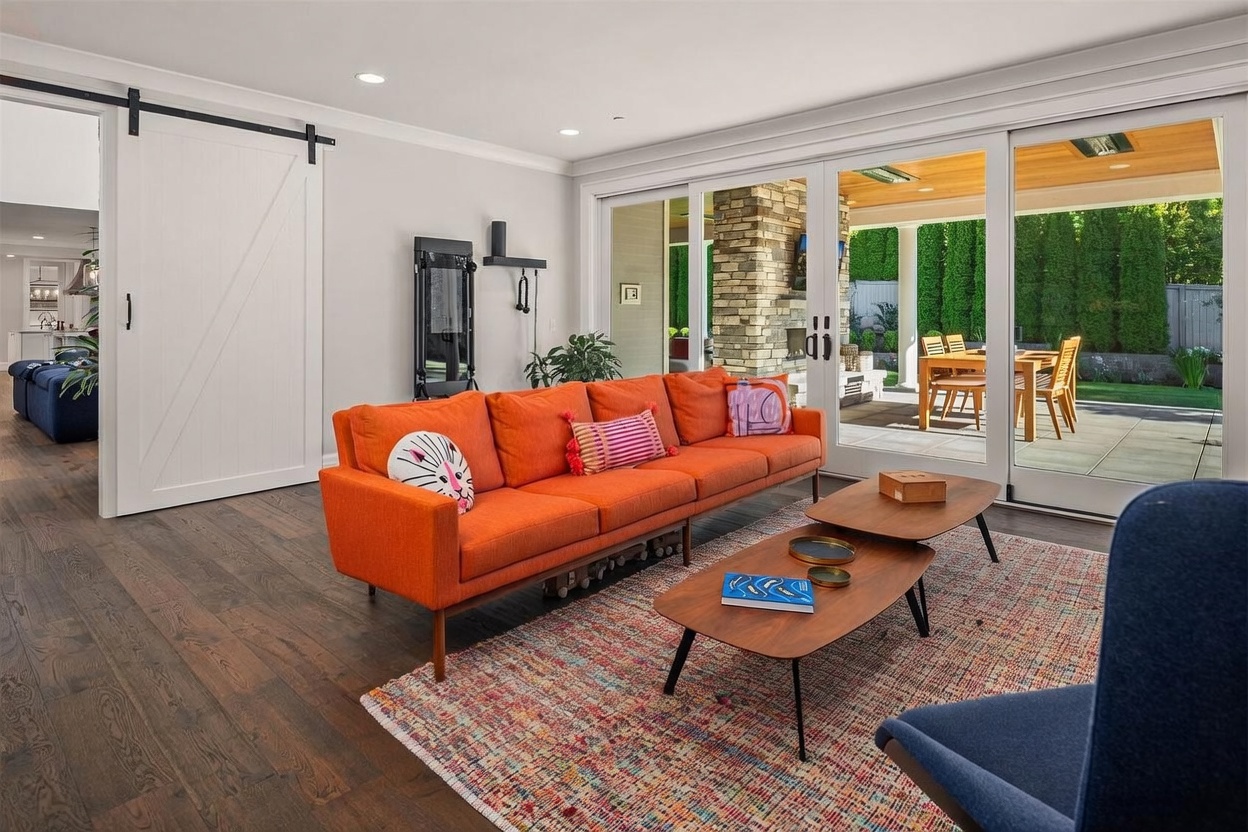 This bright living room features a vibrant orange sofa as the centerpiece, set against a backdrop of dark wood flooring and a colorful patterned area rug. Large sliding glass doors provide a seamless transition to an outdoor patio area, which includes a stone-clad chimney breast and dining space. The room is accented by a sliding barn door and modern coffee tables, creating a warm and inviting atmosphere.