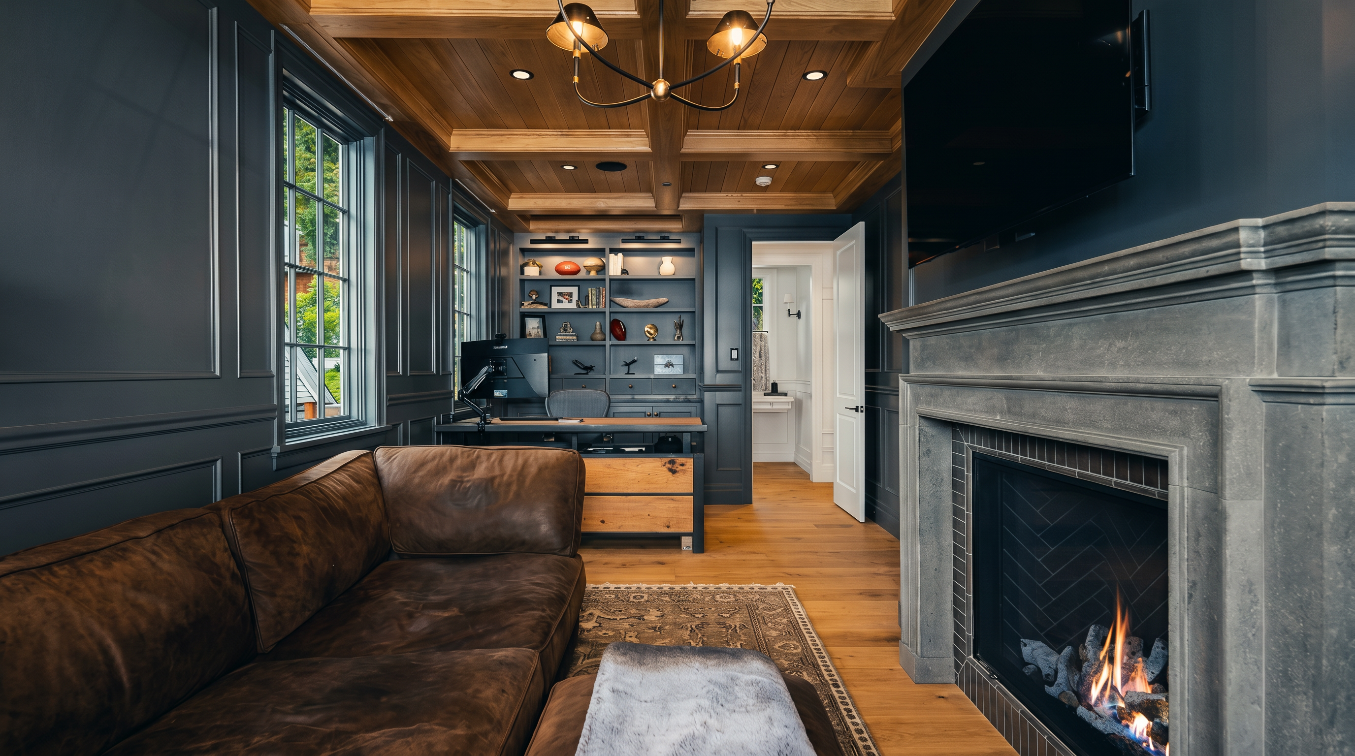 This sophisticated home office features dark, paneled walls and a coffered wood ceiling, creating a moody and professional atmosphere. A leather sofa sits on a patterned rug opposite a stone fireplace with a mounted television above, while a desk is positioned against a wall of built-in shelving. The room is illuminated by a multi-arm chandelier and natural light from the side windows, offering a refined space for work or relaxation.