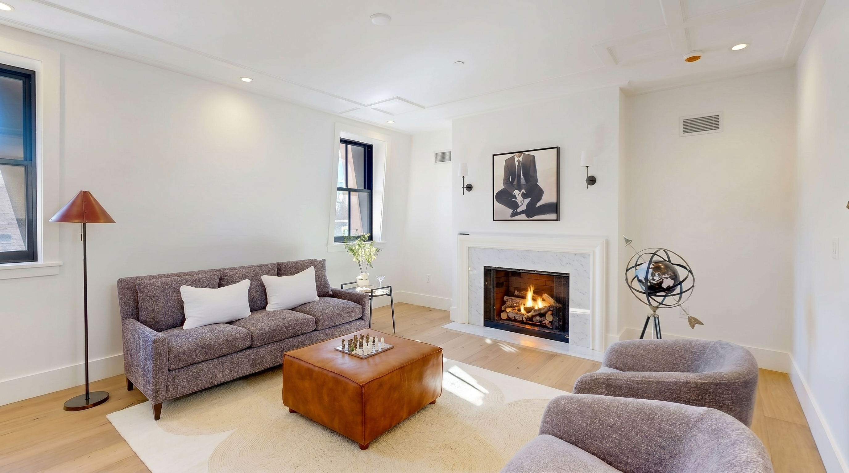 This bright and airy living room features a neutral color palette with light wood flooring and white walls. The space is anchored by a gray sofa, a large leather ottoman, and two matching armchairs, all arranged around a central fireplace with a stone surround. A decorative globe sculpture and a floor lamp add character to the room, which is illuminated by recessed lighting and natural light from the windows.