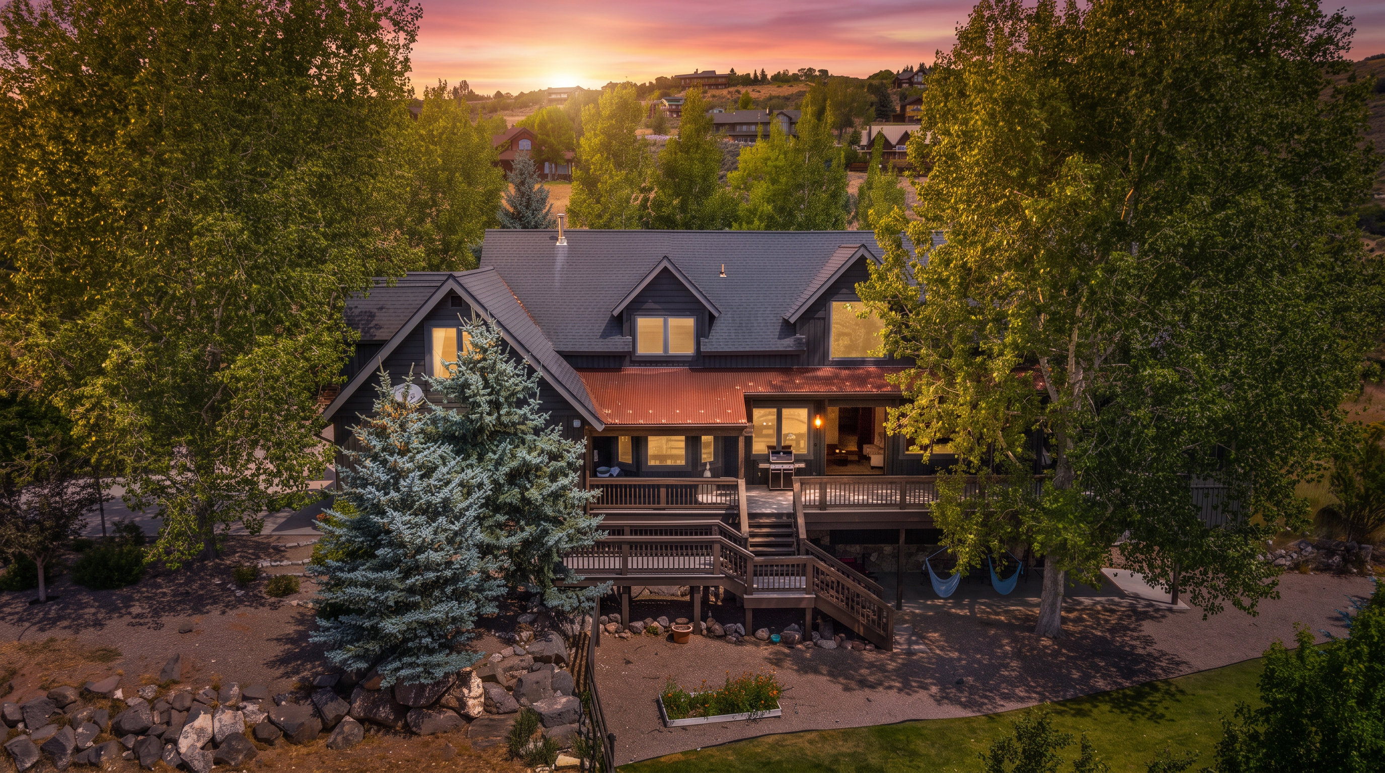 This aerial view showcases the rear of a spacious multi-story mountain-style home featuring dark wooden siding, a prominent metal roof accents, and expansive tiered decking. The property is surrounded by mature trees, a landscaped yard, and rugged rocky terrain, creating a rustic and peaceful atmosphere. The back deck provides excellent outdoor living space, perfect for enjoying the scenic, elevated hillside setting.