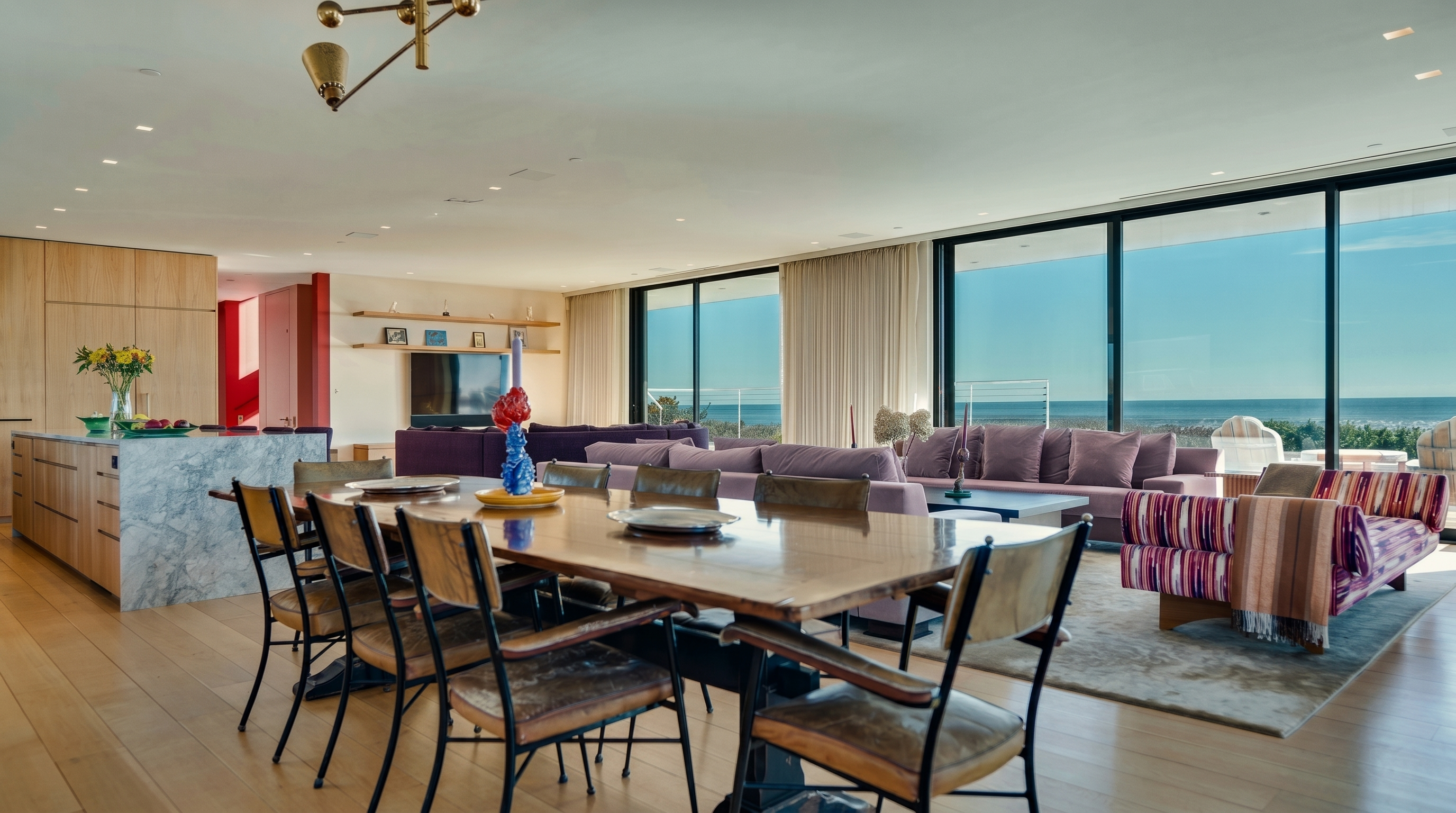 This expansive open-concept living and dining area features light wood flooring and a seamless transition to the outdoors through floor-to-ceiling glass walls. The space is anchored by a large dining table in the foreground and a comfortable purple sofa arrangement in the background, all bathed in natural light. A stone-topped kitchen island sits to the left, while the entire room offers a panoramic view of the ocean horizon.