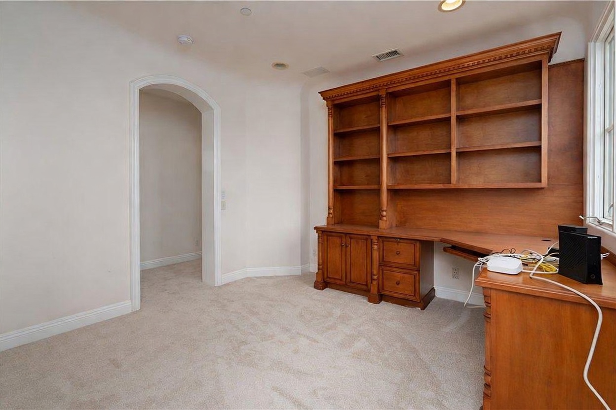 This home office features a large, built-in wooden desk and shelving unit that spans the wall, providing ample storage and workspace. The room is finished with neutral-toned walls and light-colored carpeting, creating a bright and professional atmosphere. An arched doorway leads to an adjacent space, while a window on the right allows natural light to fill the room.