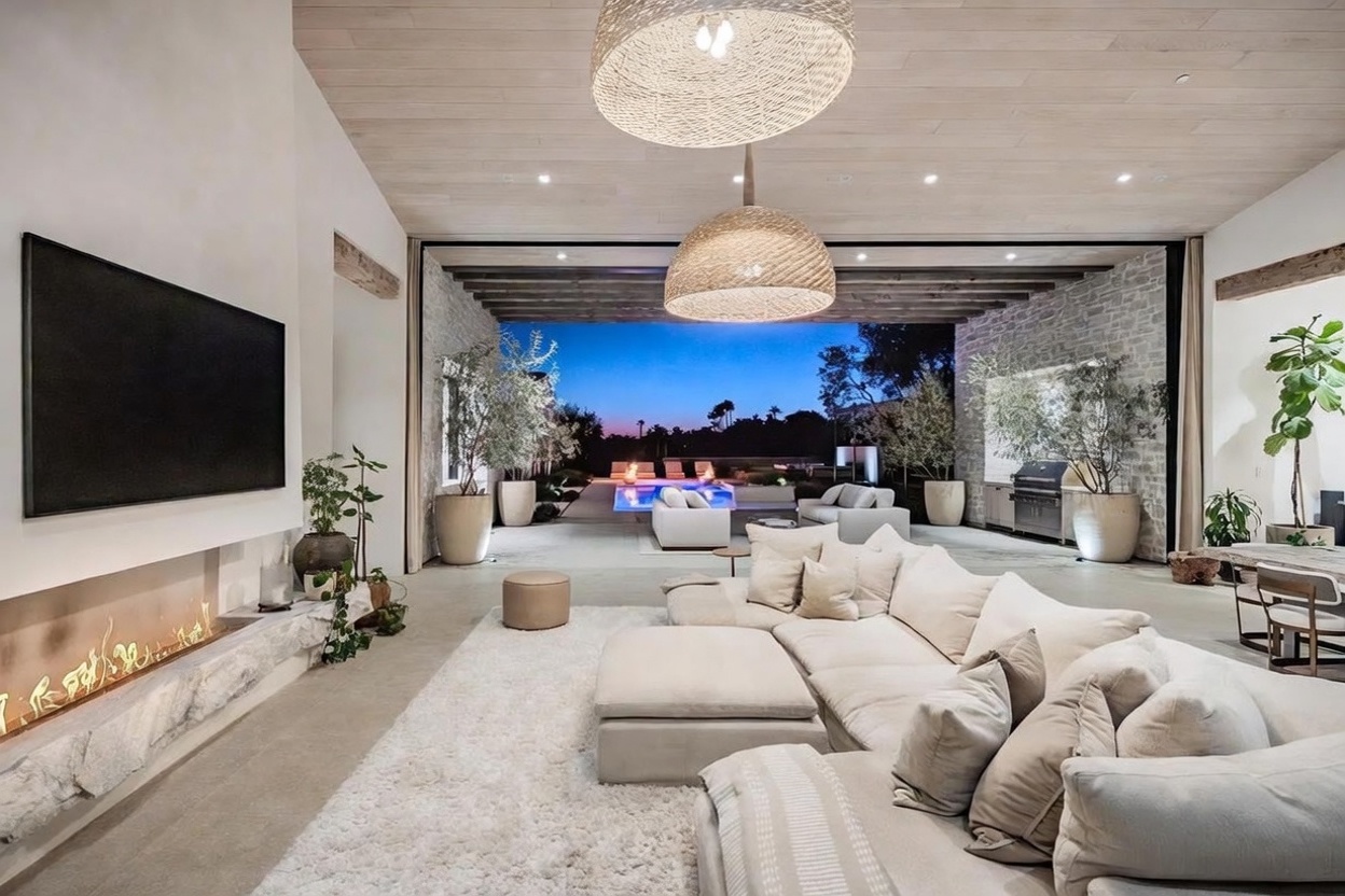 This expansive, open-concept living room features a seamless transition to an outdoor patio through a wide, retractable wall opening. The space is anchored by a large, plush sectional sofa on a textured rug, facing a linear fireplace set into a stone-clad wall beneath a wall-mounted television. Natural light wood ceilings and dual woven pendant lights enhance the airy, modern aesthetic, while the view beyond showcases a pool and landscaped yard at dusk.