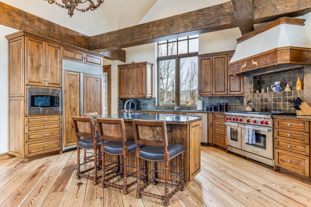 This rustic-style kitchen features warm wood cabinetry, a large central island with bar seating, and high-end stainless steel appliances, including a professional-grade range. The space is defined by exposed timber ceiling beams and light-toned wood flooring, with a large window above the sink providing natural light. The overall aesthetic is cozy and mountain-inspired, showcasing a functional layout with ample counter space and a decorative range hood.