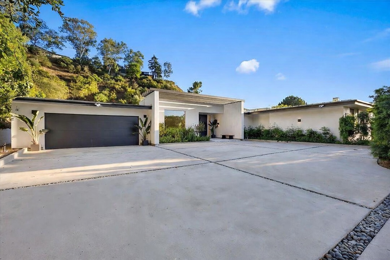 This modern single-story residence features a clean, minimalist aesthetic with a prominent dark garage door and a recessed entryway. The expansive concrete driveway provides ample parking, while the surrounding landscape includes manicured greenery and a hillside backdrop. The architectural design emphasizes horizontal lines and a bright, neutral facade, creating a sophisticated and welcoming curb appeal.