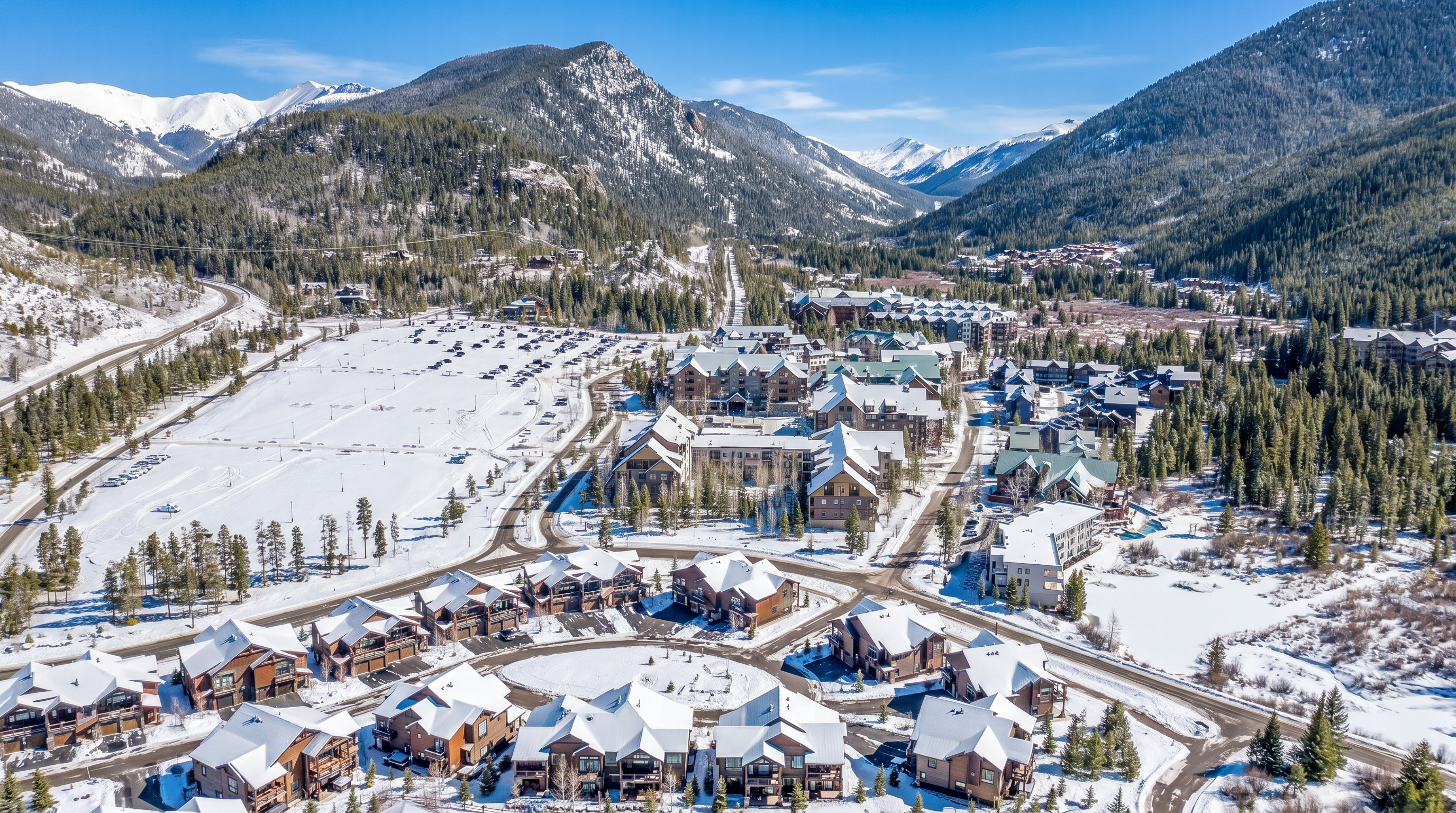 This high-angle aerial view captures a sprawling mountain resort community nestled in a lush valley surrounded by towering, snow-capped peaks. The scene features a mix of modern residential townhomes in the foreground, a large parking area, and a dense cluster of multi-story lodge-style buildings set against a backdrop of evergreen forests. The perspective emphasizes the expansive scale of the development and its seamless integration into the rugged, scenic landscape.
