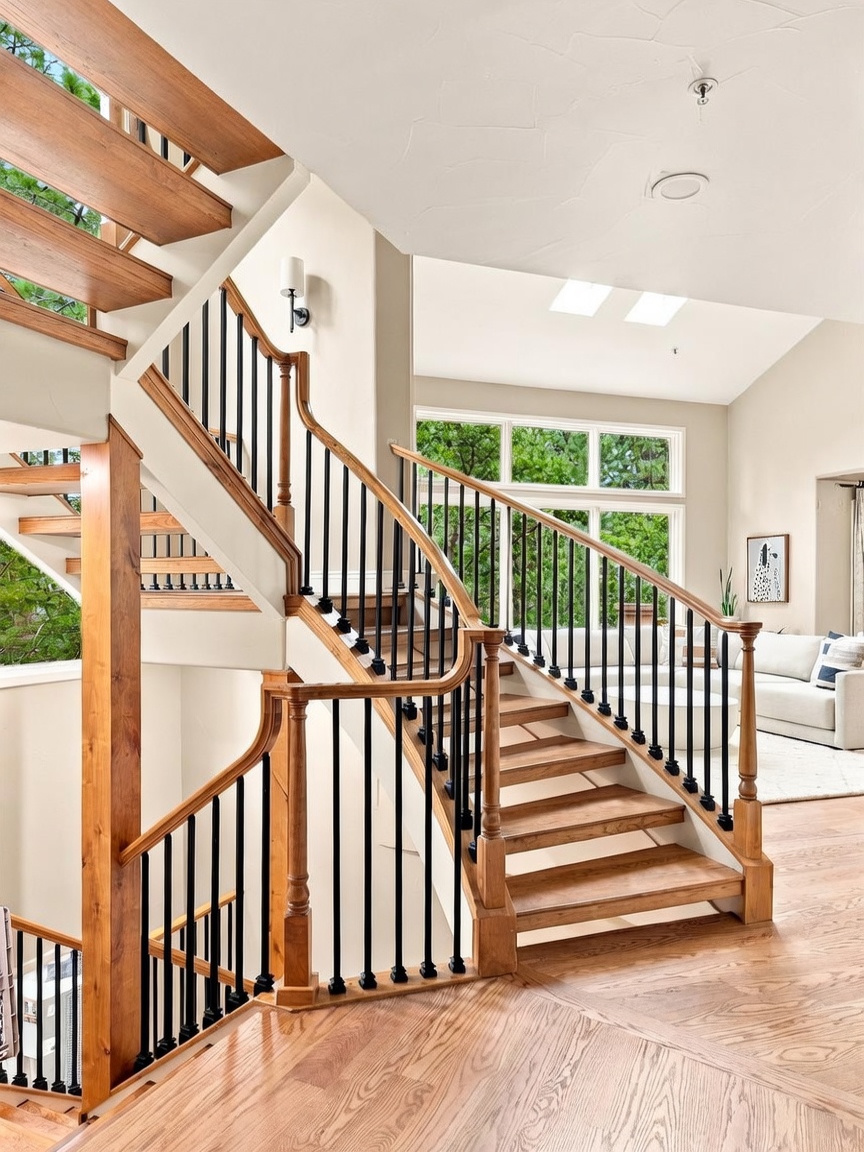 This grand entryway features a prominent staircase with light wood treads and a classic black metal balustrade. The open-concept design allows for a clear view into the adjacent living area, which is bathed in natural light from large windows. The perspective from the landing highlights the architectural flow and the bright, airy atmosphere of the home's main level.