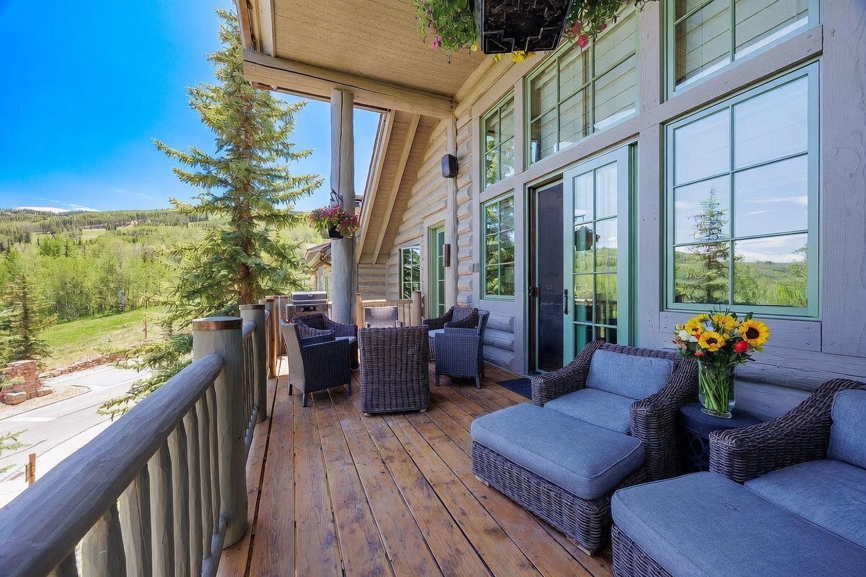 This spacious wooden deck offers a serene outdoor living space with comfortable wicker seating and a vibrant floral arrangement. The area overlooks a lush, tree-lined landscape, providing a peaceful mountain retreat atmosphere. Large windows and glass doors connect the interior to the deck, enhancing the home's indoor-outdoor flow.