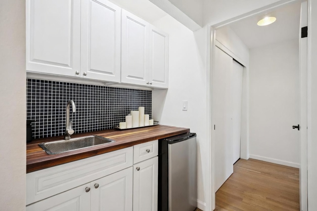 This compact kitchenette features white shaker-style cabinetry paired with a dark wood-toned countertop and a striking black grid-patterned tile backsplash. A stainless steel sink with a modern gooseneck faucet sits beside a small under-counter refrigerator, creating a functional wet bar or auxiliary prep area. The space is bright and clean, transitioning into a hallway with light-toned wood flooring and white walls.